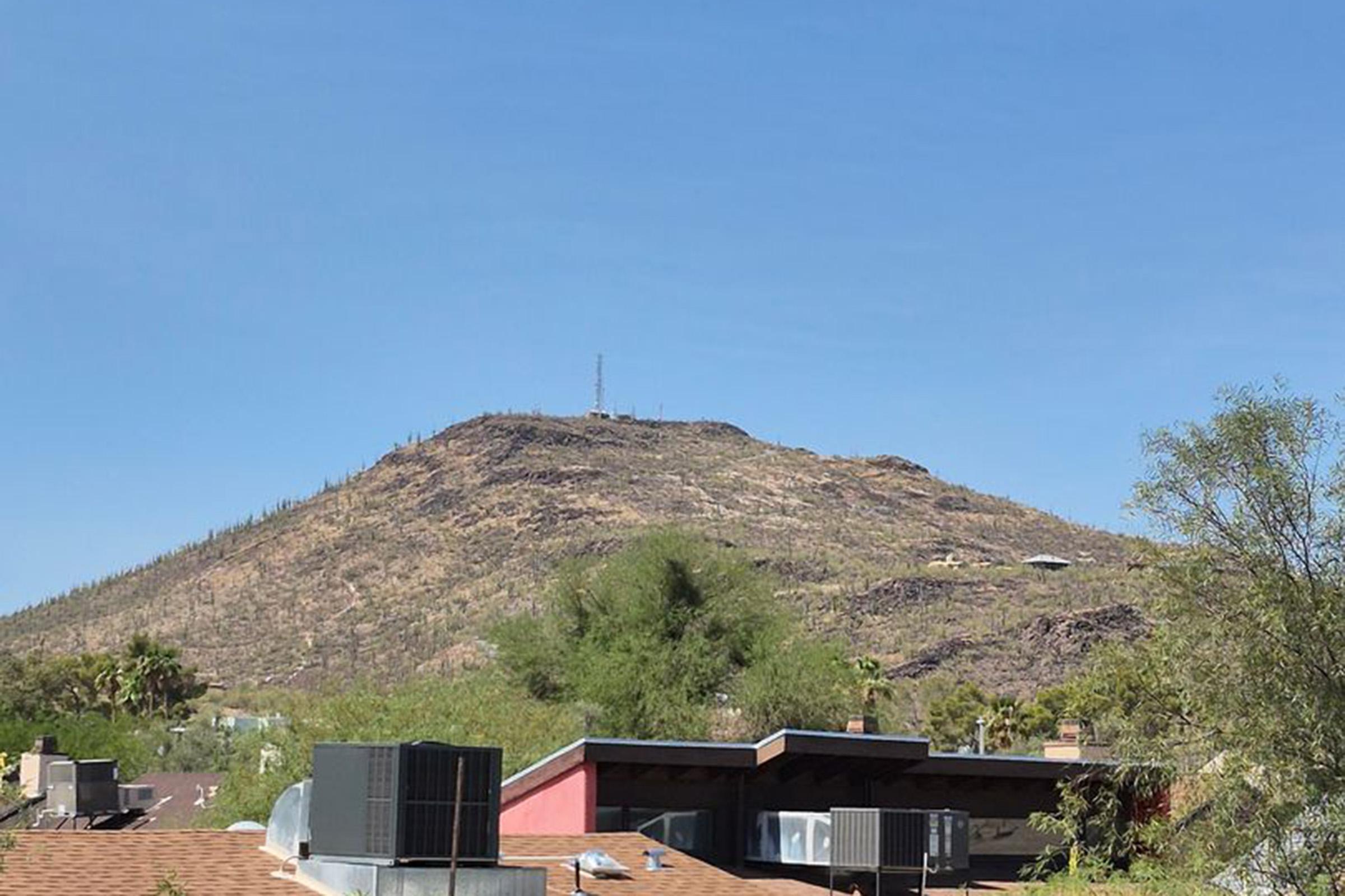 A dry mountain landscape under a clear blue sky, featuring a hillside with sparse vegetation and rocky outcrops. In the foreground, there are rooftops with air conditioning units, contrasting with the natural terrain behind. A communication tower is visible on the mountain peak.