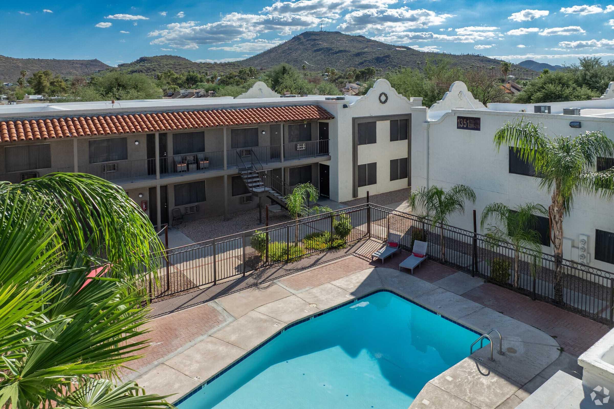 A view of a residential complex featuring a swimming pool surrounded by palm trees, with two-story buildings in the background. The scene includes mountain hills under a partly cloudy sky, showcasing a tranquil outdoor environment. The area is well-maintained with landscaping and a clear pool.