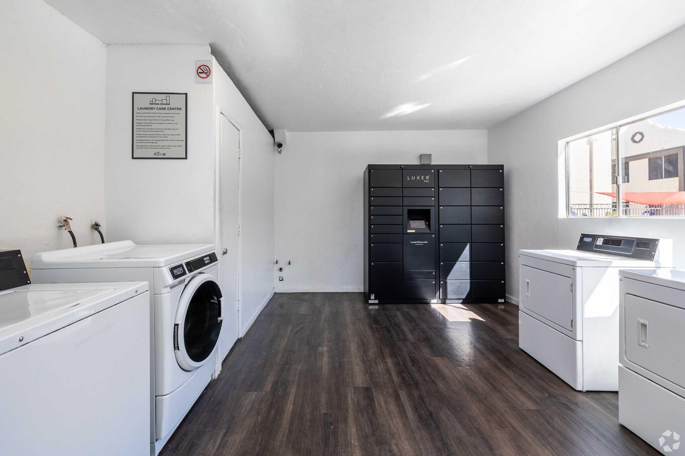 A bright laundry room featuring two white washing machines and one dryer, with a modern black package locker against the wall. The space has light-colored walls and a clean, open layout, illuminated by natural light coming through a window. A sign is mounted on the wall near the entrance.