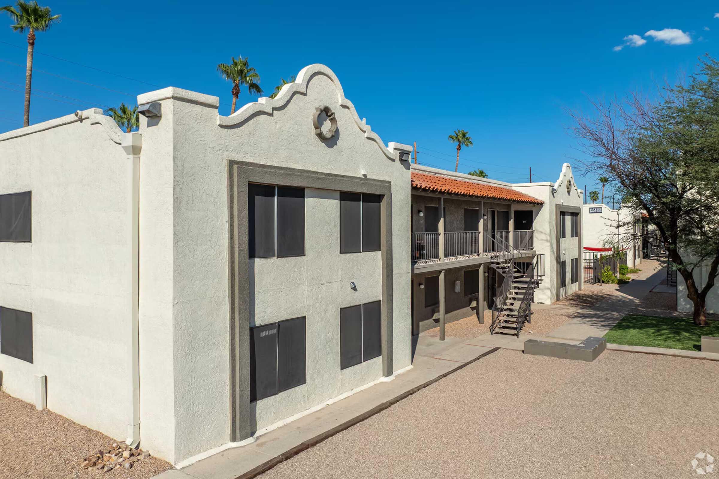 Two-story white apartment building with a decorative, curved roofline and balcony, set against a clear blue sky with palm trees in the background. A staircase leads to the second floor, and the surrounding area features gravel landscaping and green plants.