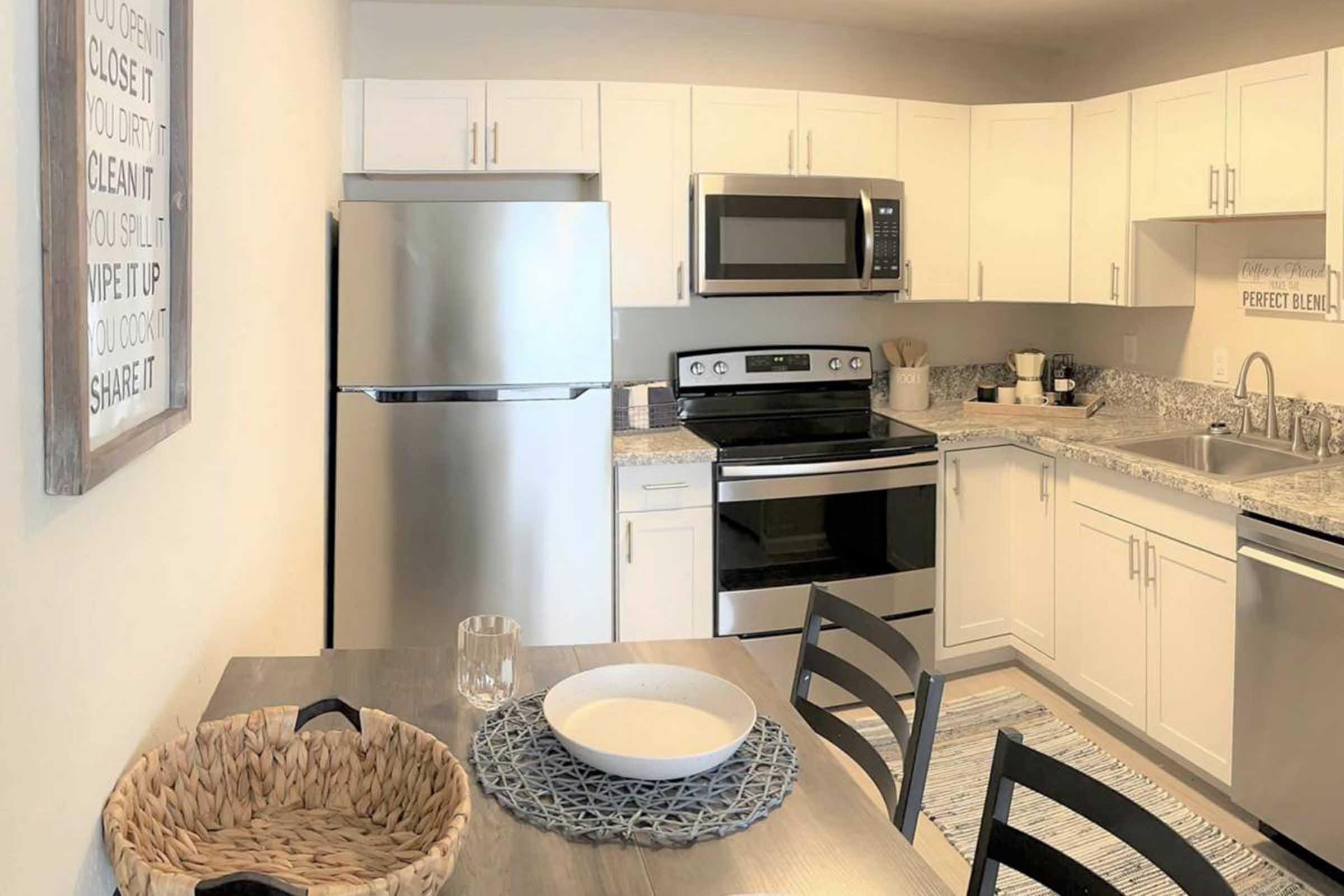 A modern kitchen featuring white cabinetry, stainless steel appliances including a refrigerator and microwave, and a sleek countertop. A dining table with a woven basket and a decorative bowl sits nearby. The space is bright and well-organized, emphasizing a clean, contemporary design.