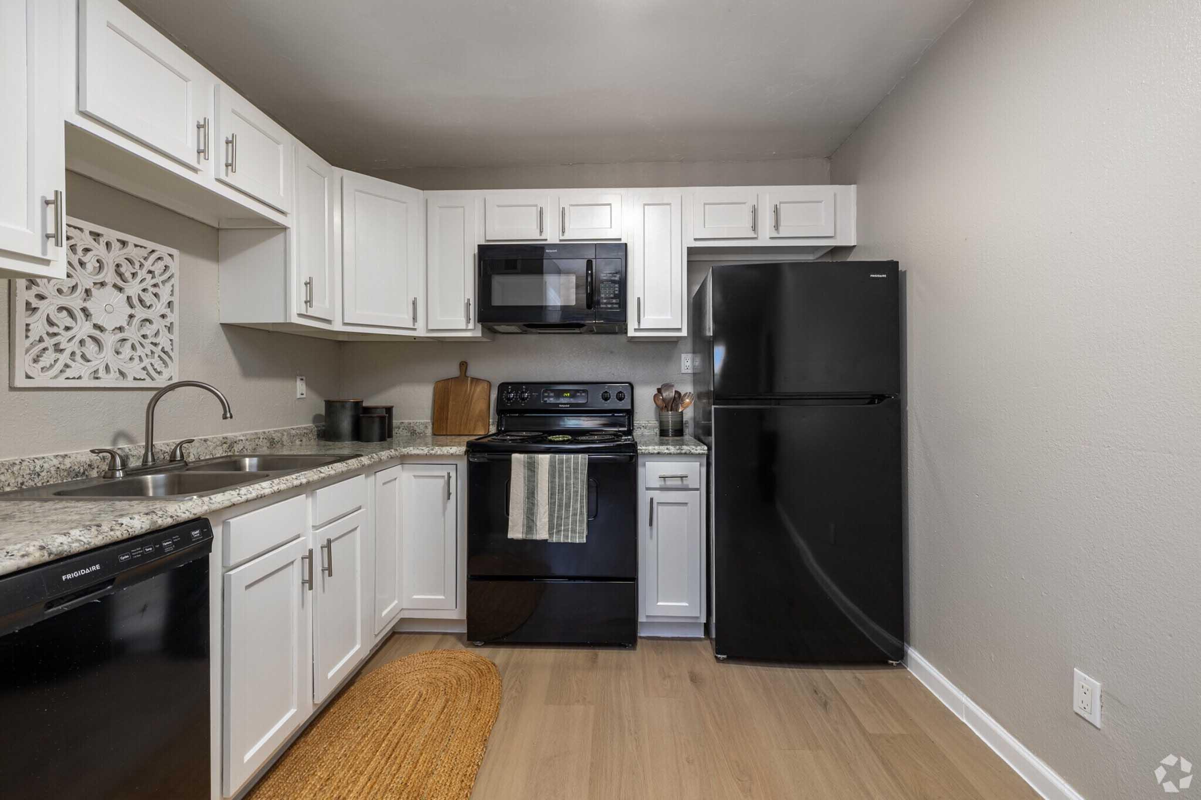 Modern kitchen featuring white cabinetry, a black refrigerator, and a black stove. The countertop has a decorative piece and there is a stainless steel sink. A dishwasher is installed, and the flooring is light-colored wood. A round woven rug is placed under the sink area.
