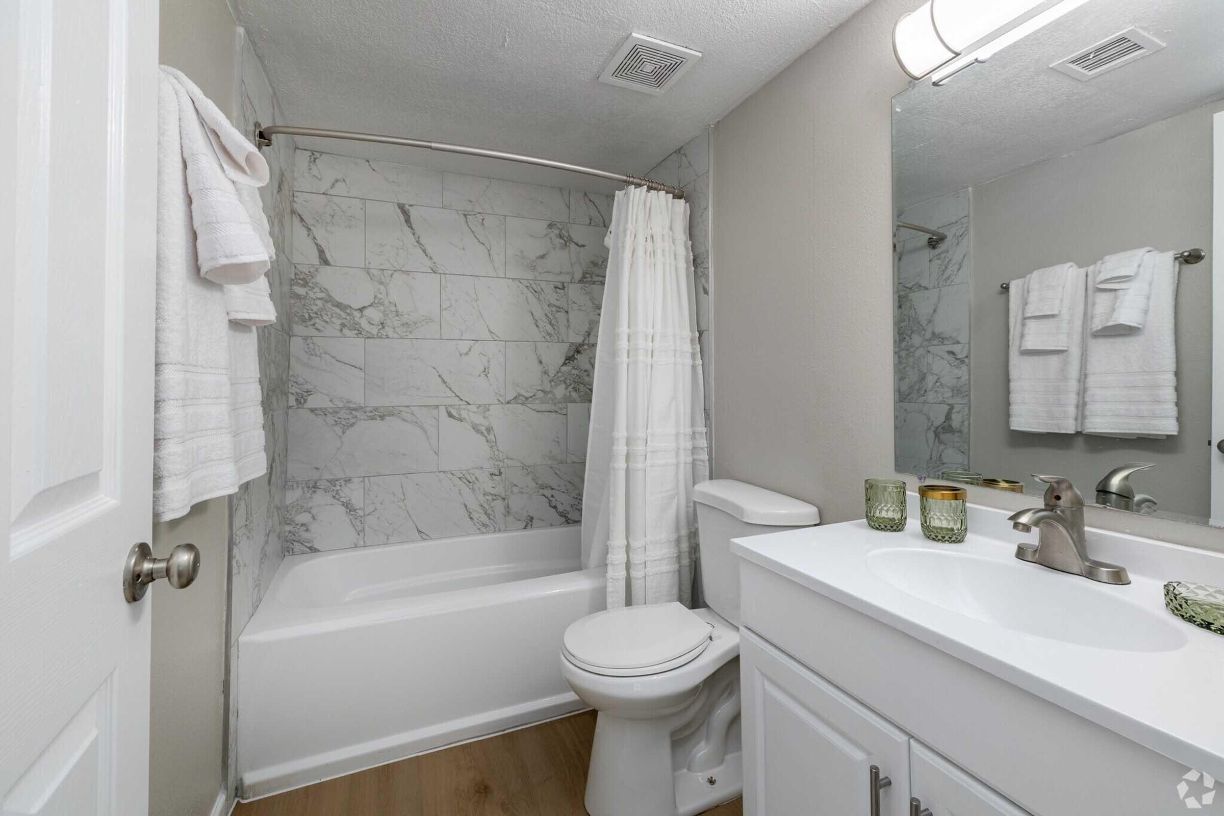 A modern bathroom featuring a white bathtub with a curved shower rod and a white shower curtain. The walls are adorned with marble-like tiles. A white vanity with a sink and faucet is on one side, accompanied by neatly arranged towels and decorative candles. Bright lighting enhances the clean, minimalistic design.