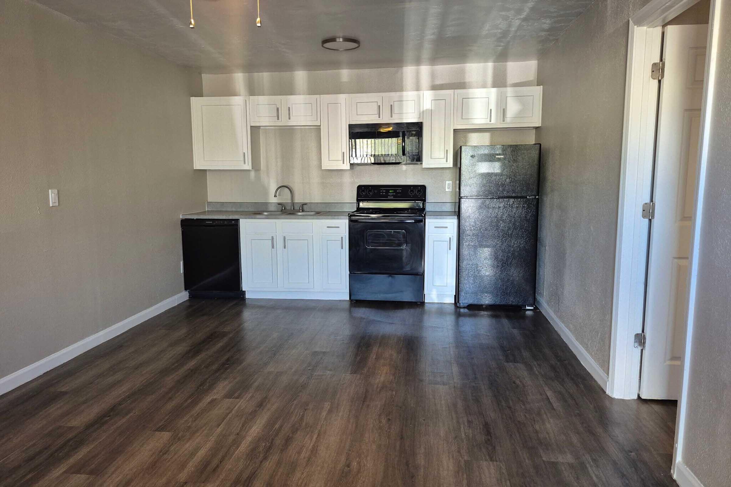 A clean, modern kitchen featuring white cabinetry, a black stove, a stainless steel refrigerator, and a black dishwasher. The kitchen has a light gray wall color and wooden floors, creating a spacious and inviting atmosphere. Natural light comes through a nearby window.
