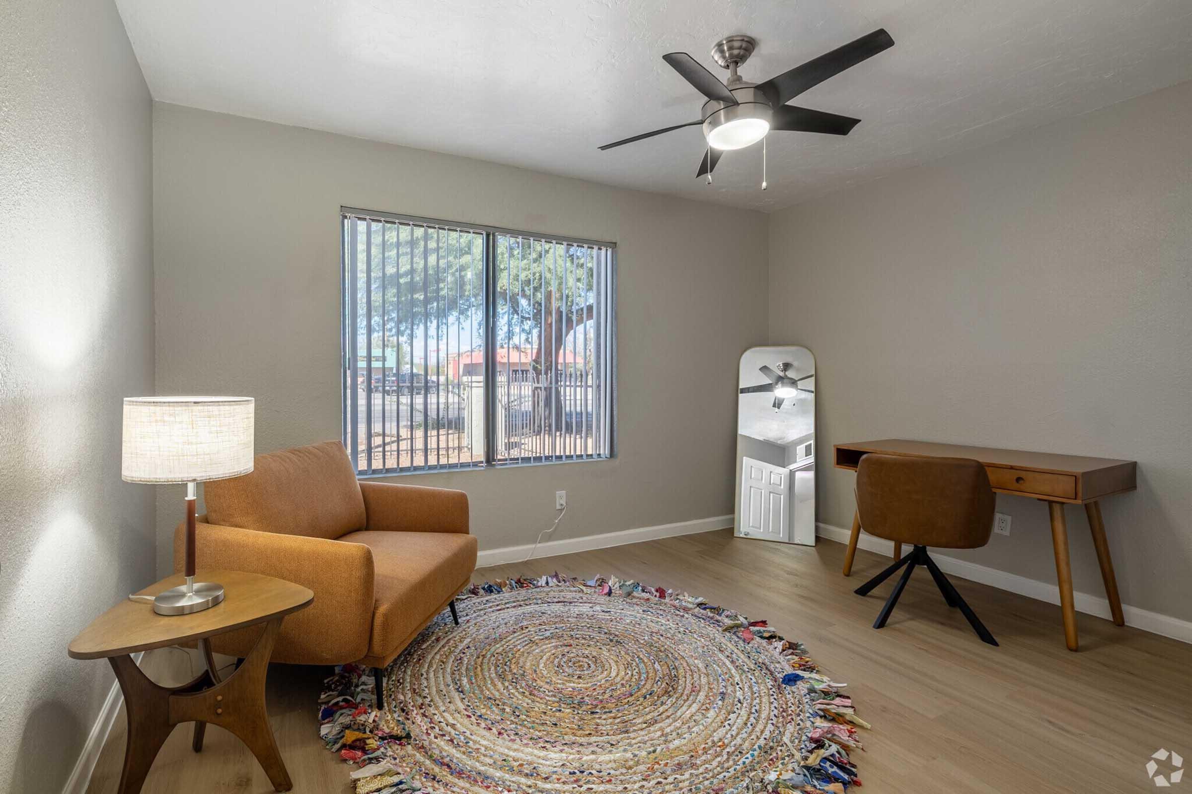 A cozy living space featuring a light brown armchair, a round area rug, a wooden desk with a chair, and a full-length mirror. Natural light filters in through a window with vertical blinds, providing a view of the outdoors. A modern ceiling fan adds to the room's comfortable ambiance.