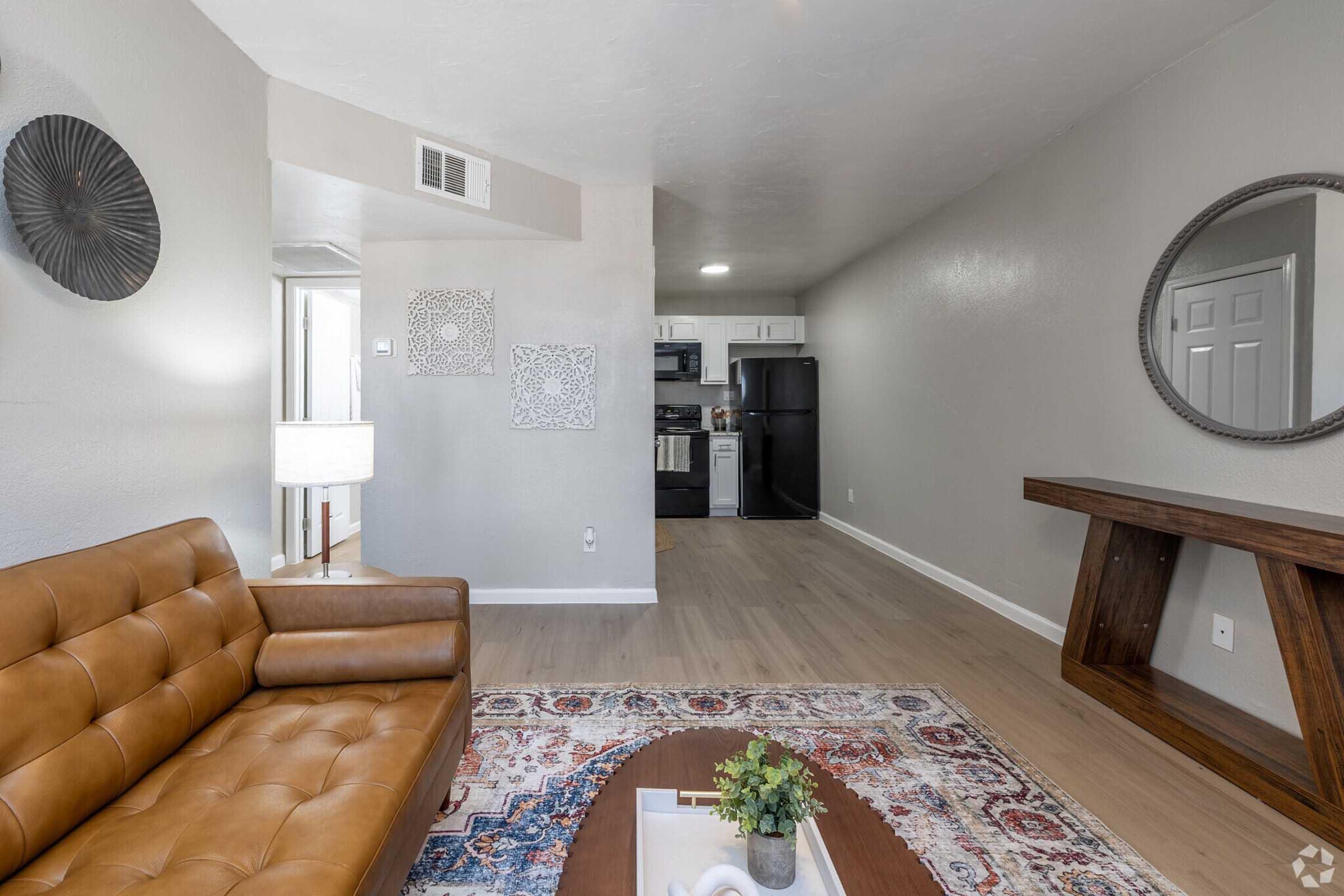 A modern living area featuring a brown leather sofa, a coffee table with decorative items, and a rug. The open space connects to a kitchen visible in the background, equipped with appliances. A wall-mounted mirror and framed art pieces add a touch of style to the room. Soft lighting enhances the inviting atmosphere.