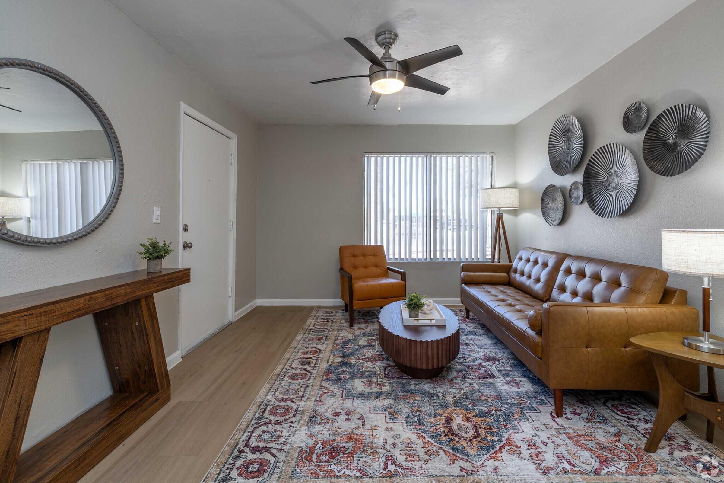 A modern living room featuring a brown leather sofa, an armchair, a round wooden coffee table, and a decorative rug. The walls are adorned with metal wall art, and there's a round mirror above a wooden console table. Natural light streams in through window blinds, creating a warm, inviting atmosphere.
