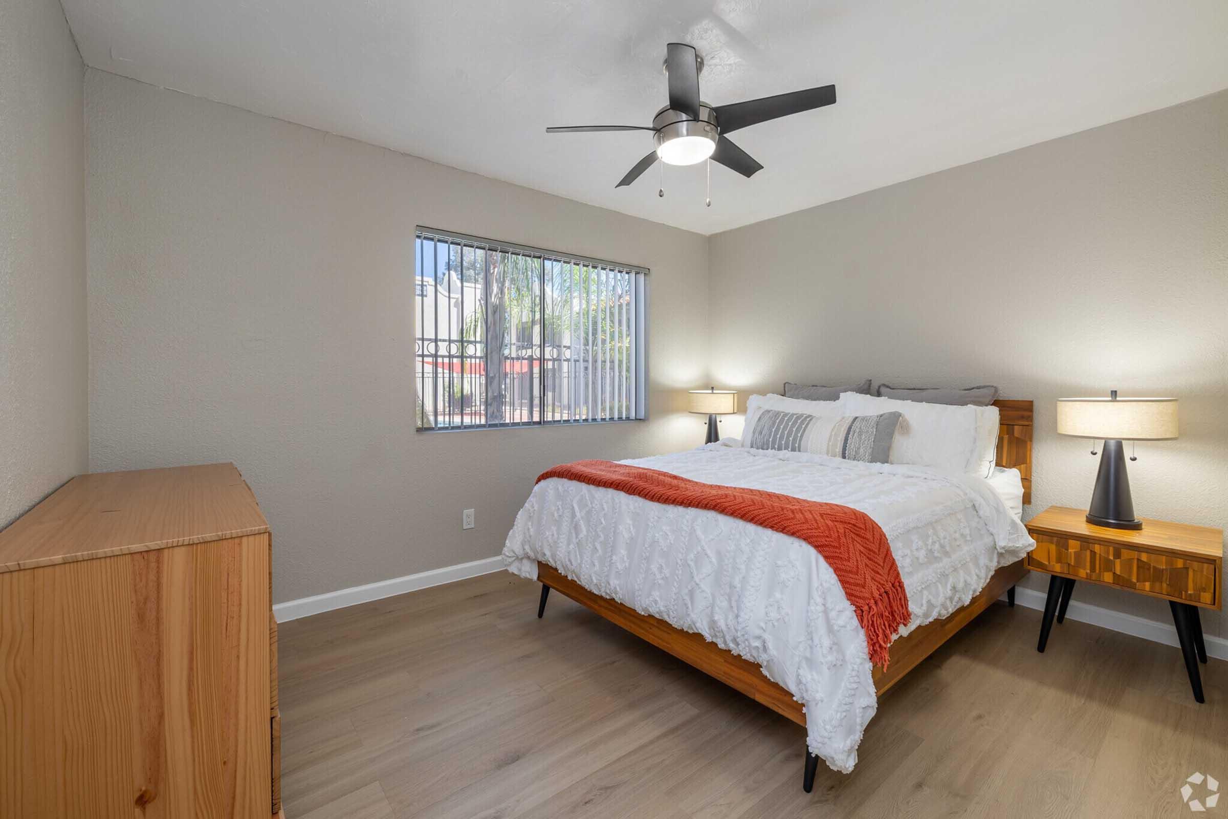 Bright bedroom with a large bed featuring white bedding and decorative pillows. A wooden dresser is against one wall, and a bedside table with a lamp sits on either side of the bed. A ceiling fan is visible, and a window with vertical blinds allows natural light to enter the room. Wooden flooring completes the cozy aesthetic.
