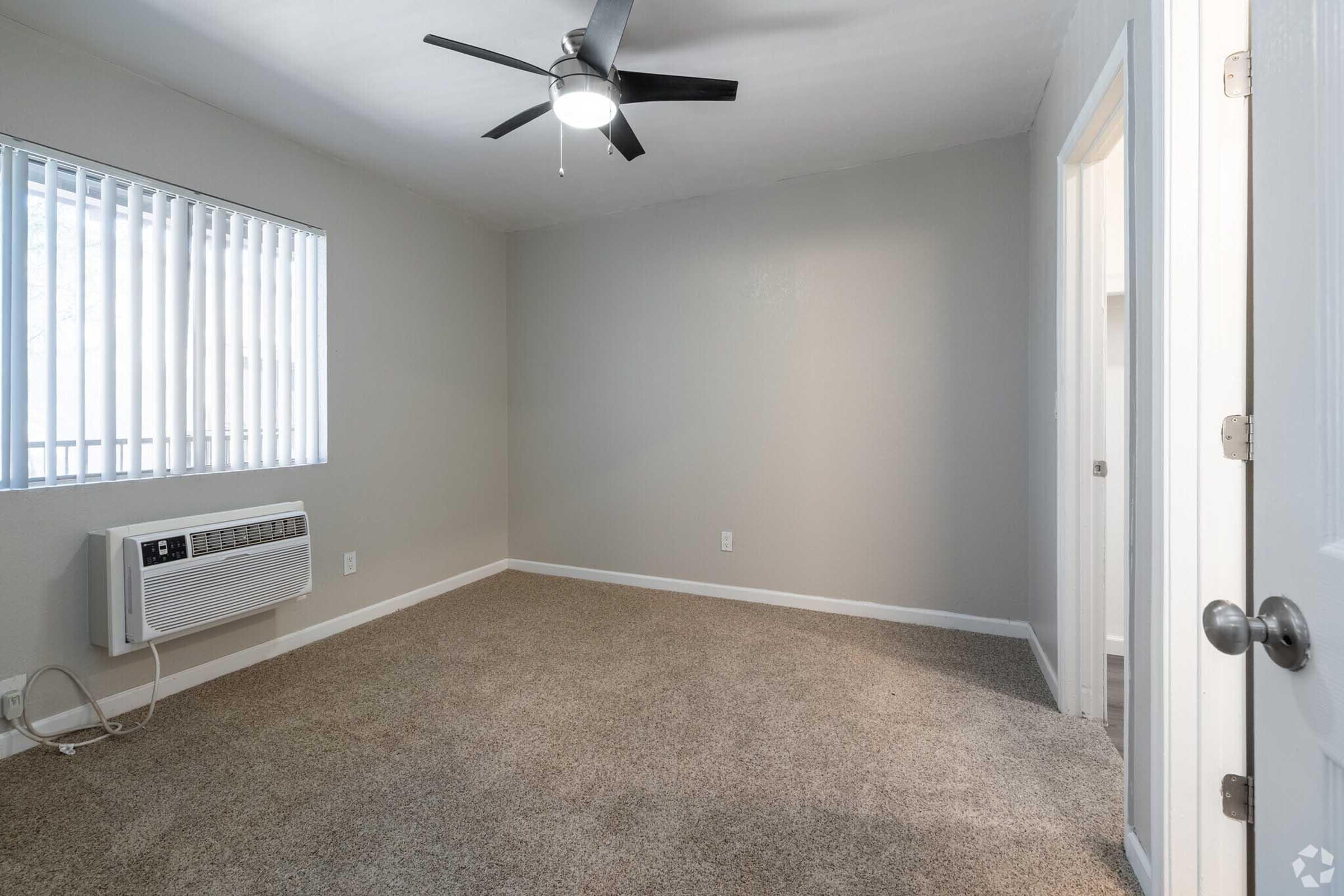 Empty room with beige carpet, a ceiling fan, and a wall-mounted air conditioning unit. There are white blinds on the window, and two doors leading to other areas. The walls are painted light gray, creating a simple and clean aesthetic.