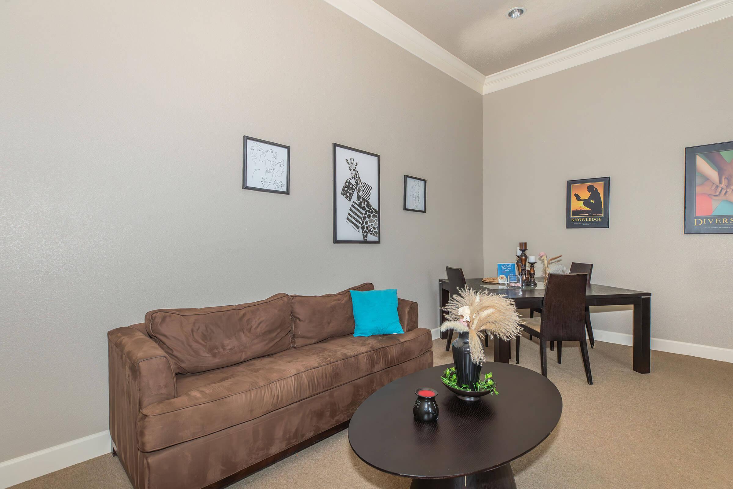 A cozy living room featuring a brown couch with a light blue cushion, a black coffee table with decorative items, and a dining area with a dark table. The walls are adorned with framed artwork, adding a touch of personality to the space. The room has neutral tones and soft lighting, creating a welcoming atmosphere.