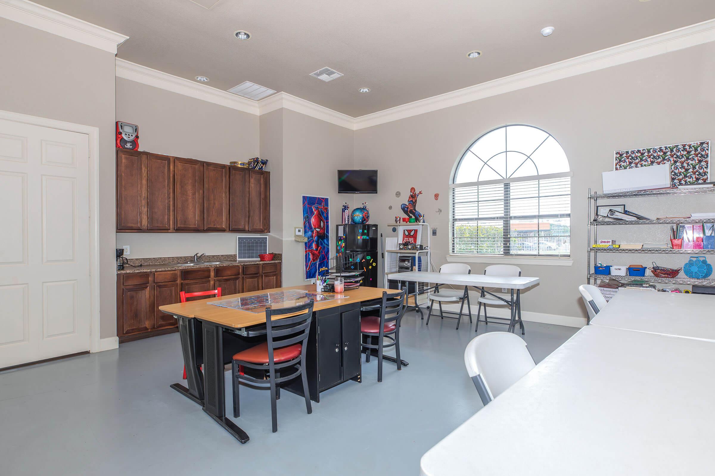 A spacious room with light gray flooring, featuring a large arched window. On one side, there are wooden cabinets and a countertop with an appliance, while the other side has multiple tables and chairs set up for activities. Shelves display various colorful objects and decorations.