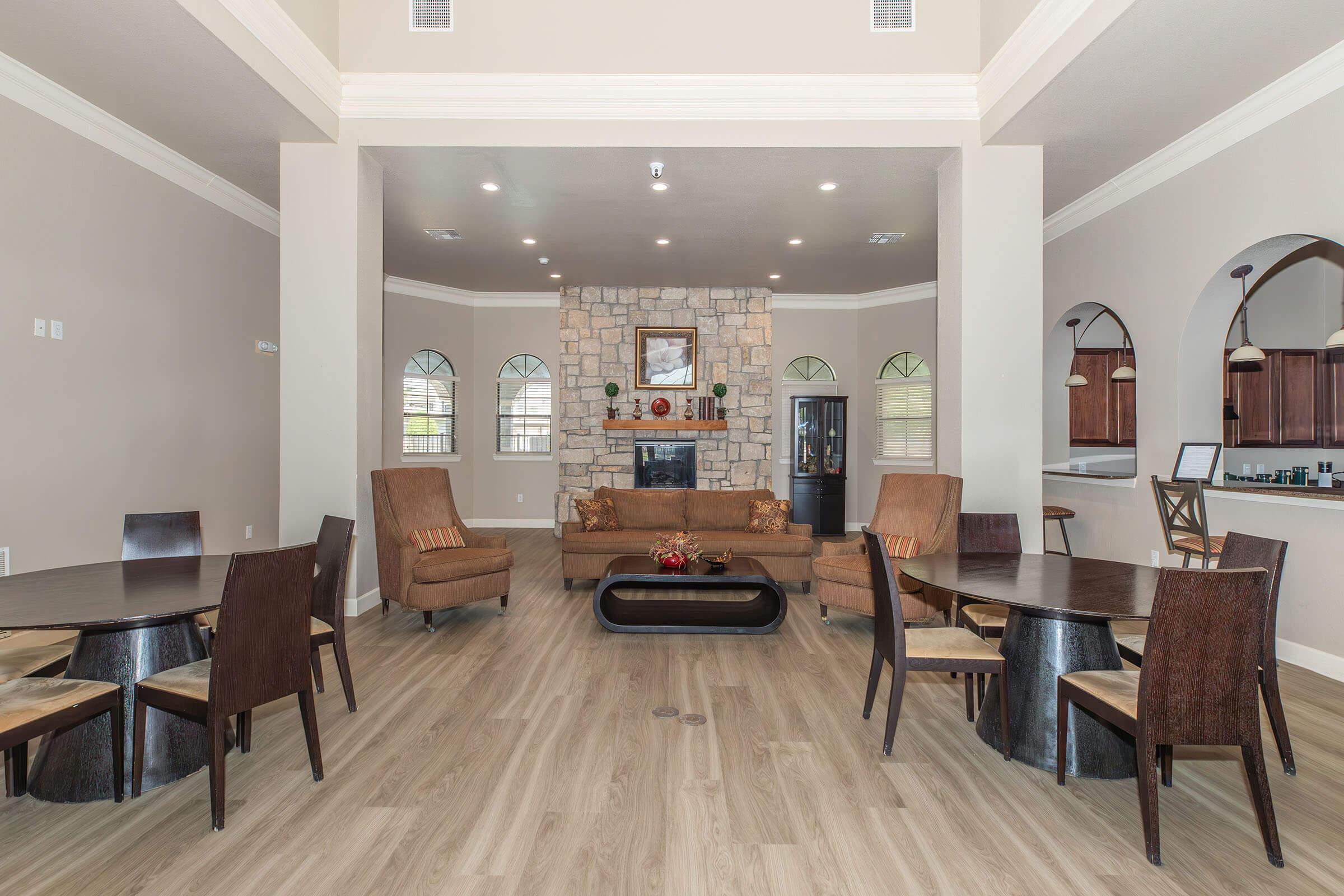 Interior of a spacious living area featuring a stone fireplace at the center, wooden flooring, and large windows. The room includes two seating areas with brown chairs and a wooden coffee table, as well as multiple dining tables arranged around the space. Soft, neutral colors create a welcoming atmosphere.