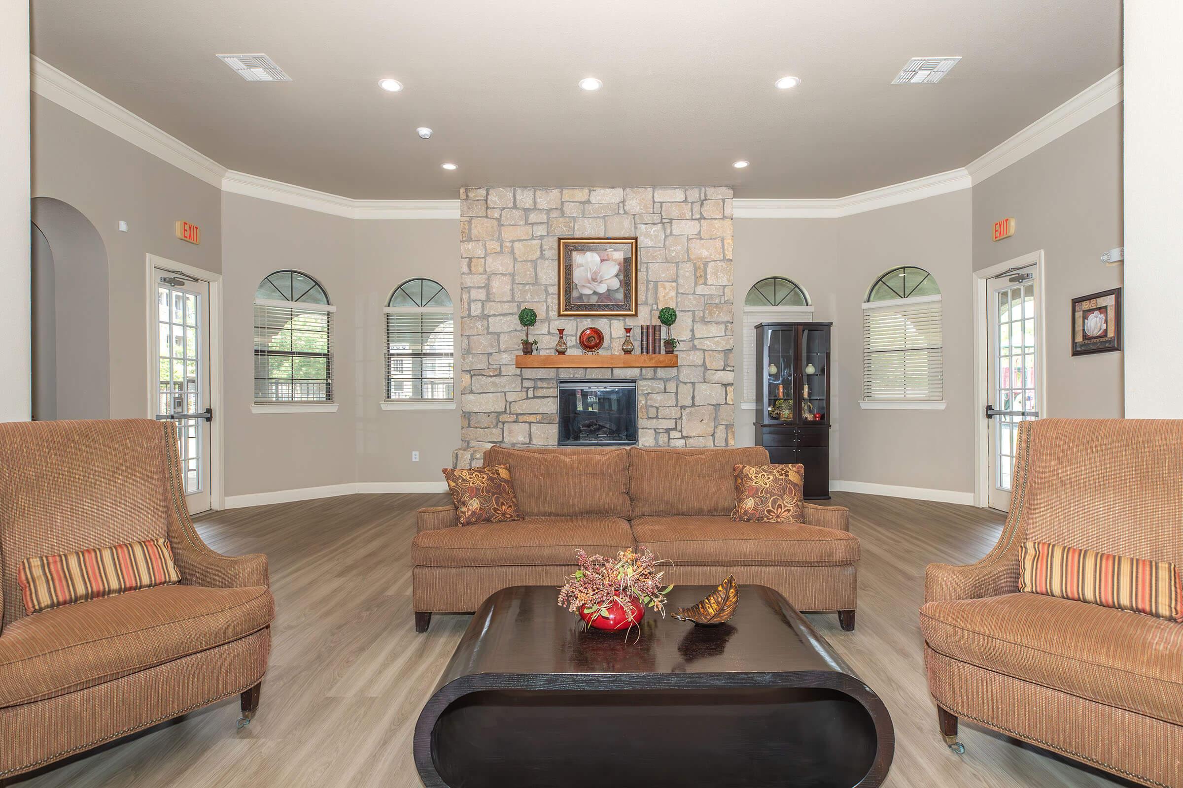 A cozy living room featuring a stone fireplace, a wooden coffee table, and two armchairs with decorative pillows. The walls are painted a light color, and there are large windows with arched tops allowing natural light. A piece of artwork is displayed above the fireplace, adding to the room's inviting atmosphere.