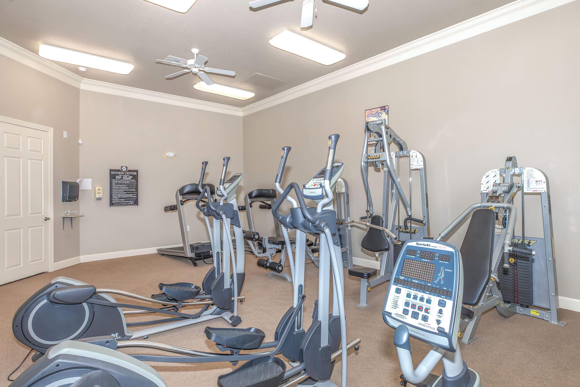 A well-lit gym interior featuring multiple pieces of exercise equipment, including elliptical machines, treadmills, and strength training machines. The walls are painted in a neutral color, and there are ceiling fans for ventilation. A door is visible in the background along with a bulletin board.