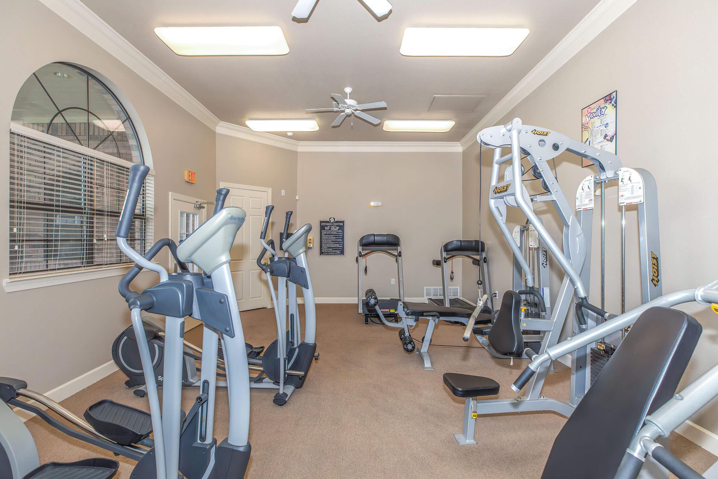 A well-lit gym room featuring various exercise equipment, including cardio machines, weight benches, and multi-purpose gym stations. The walls are painted in neutral colors, and a large window provides natural light. Ceiling fans and overhead lights enhance the space's comfort and brightness.