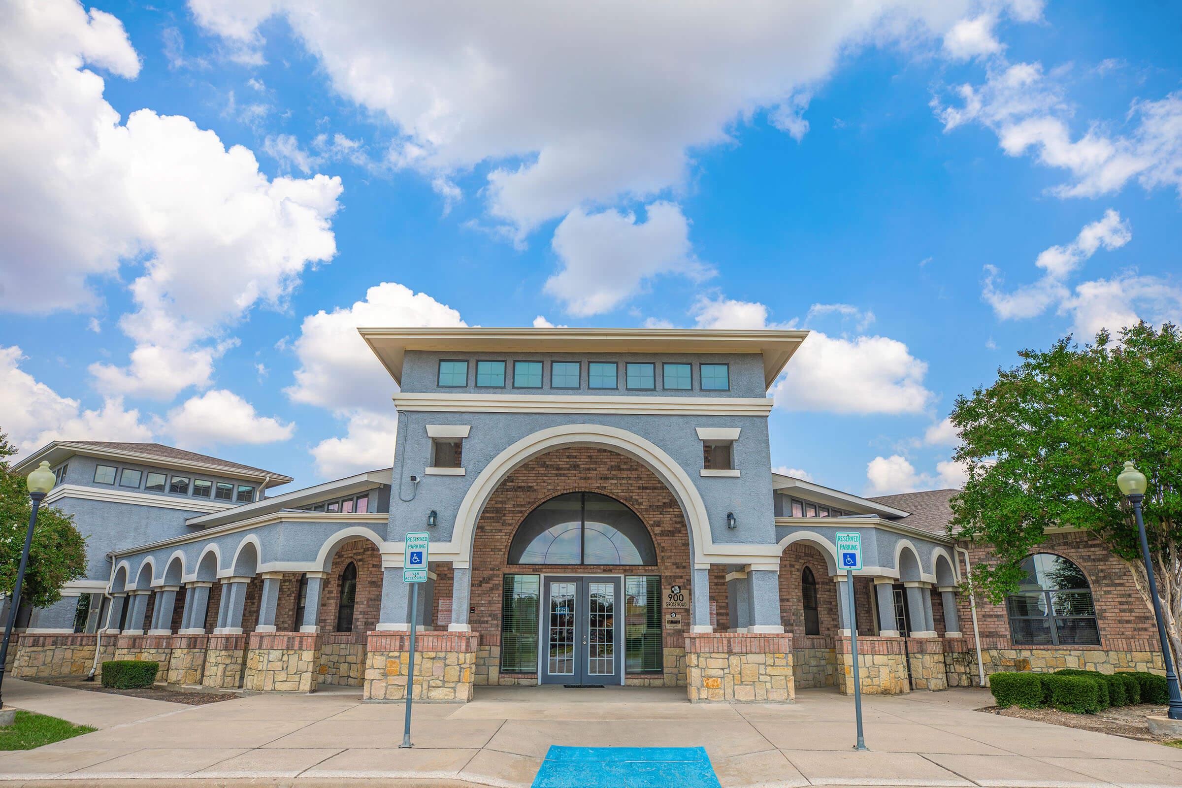 A modern building featuring a stone facade and large arched windows, set against a blue sky with fluffy clouds. The entrance is marked by double doors and is flanked by trees. Sidewalks and accessible parking spaces are visible in front of the structure, indicating a welcoming environment.