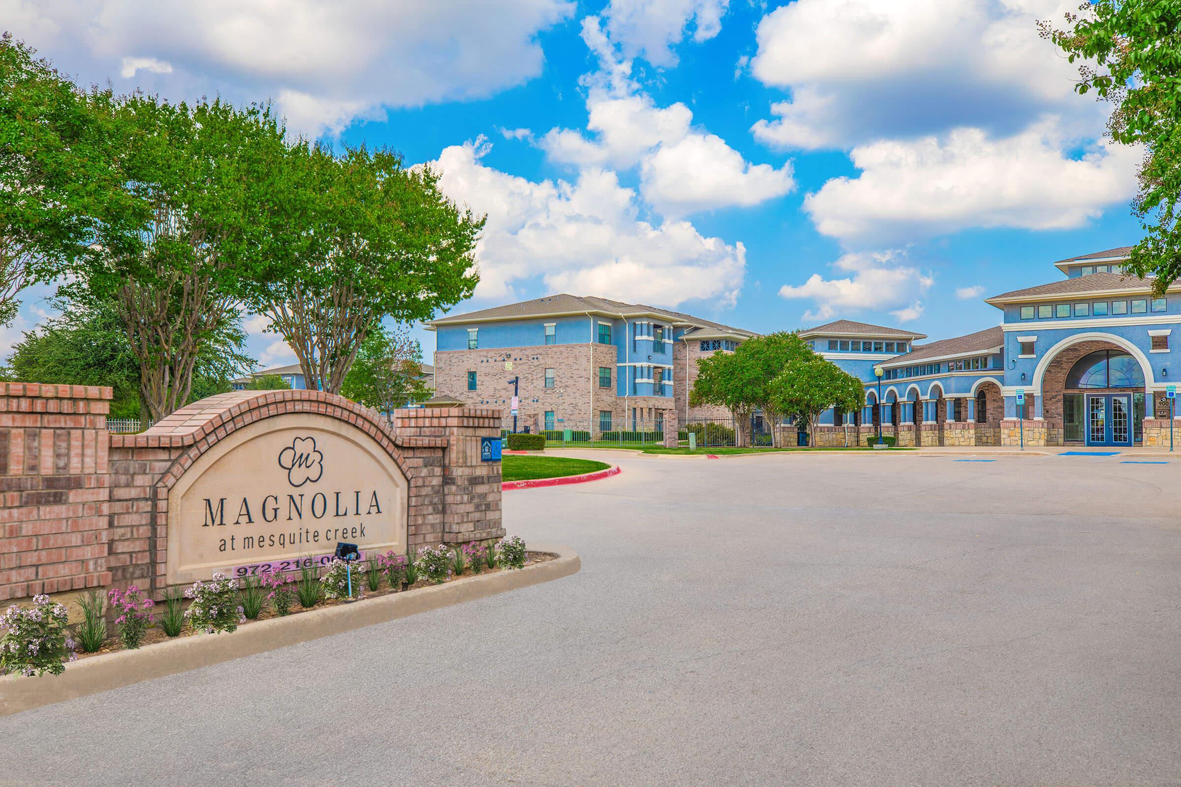 A brick sign featuring the name "Magnolia at Mesquite Creek" is prominently displayed in front of a multi-story residential building. The building has a mix of stone and blue siding, surrounded by green trees and shrubs, with a clear blue sky and fluffy clouds in the background.