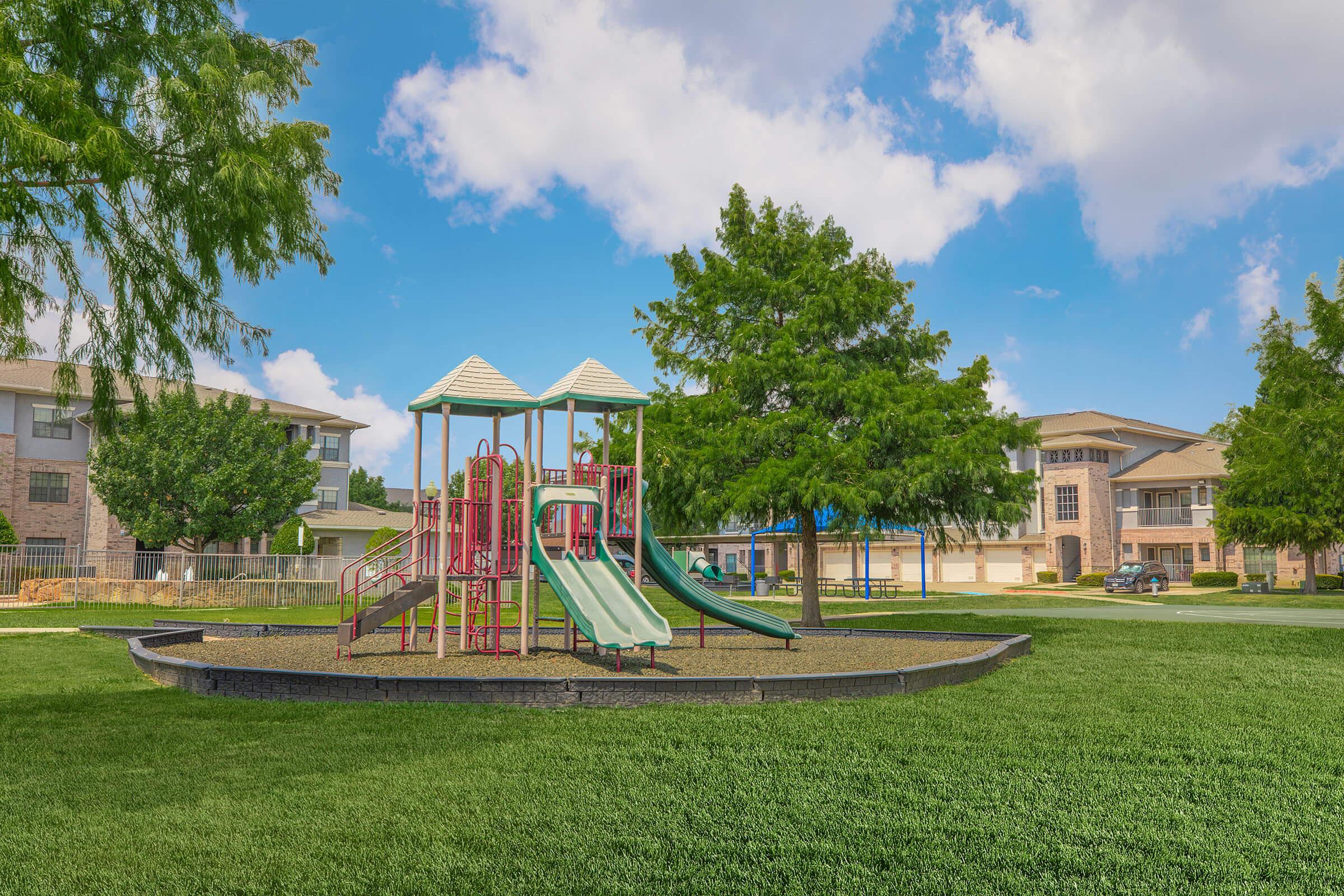 A colorful playground with two slides and a climbing structure, surrounded by green grass. In the background, there are residential buildings and trees under a blue sky with white clouds. The setting is bright and inviting, perfect for children to play.