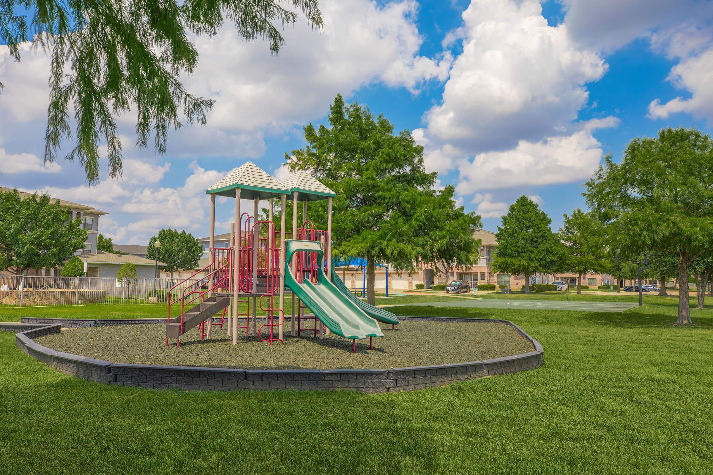 A colorful playground featuring two tall slides and a climbing structure, located on a grassy area surrounded by trees. The sky is partly cloudy, and there are residential buildings in the background. The scene conveys a cheerful outdoor space for children to play.