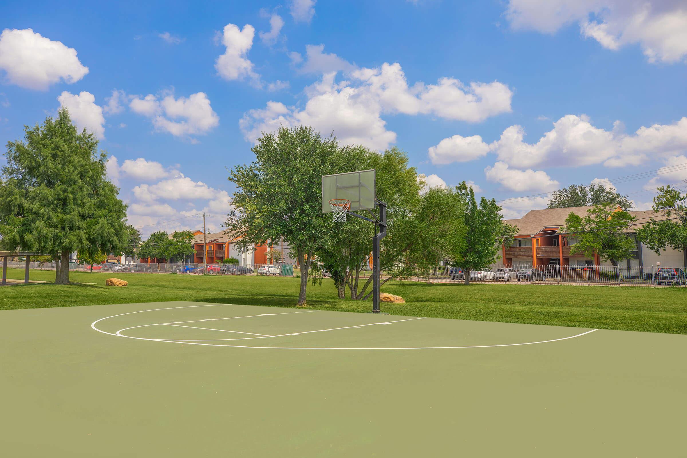 A basketball court with a hoop set against a backdrop of blue sky and fluffy clouds. Surrounding the court are green trees and a grassy area, with residential buildings visible in the distance. The scene is bright and inviting, ideal for outdoor sports and recreation.