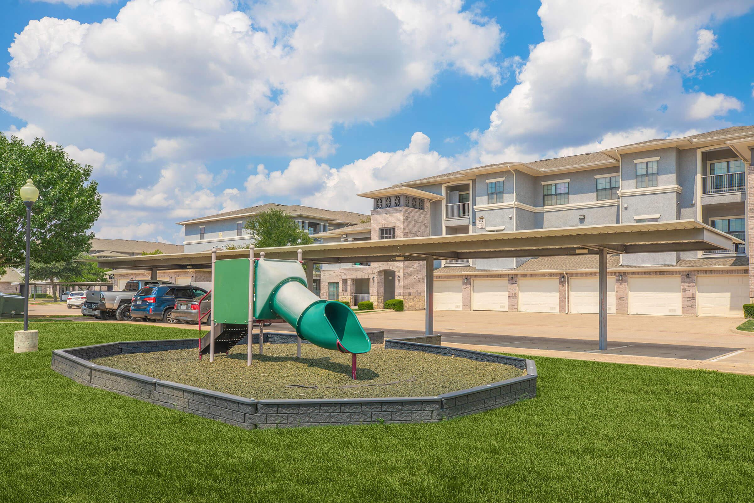 Playground area featuring a green slide on a bed of gravel, surrounded by grassy landscaping. In the background, an apartment complex with multiple stories and covered parking spaces is visible under a partly cloudy sky.