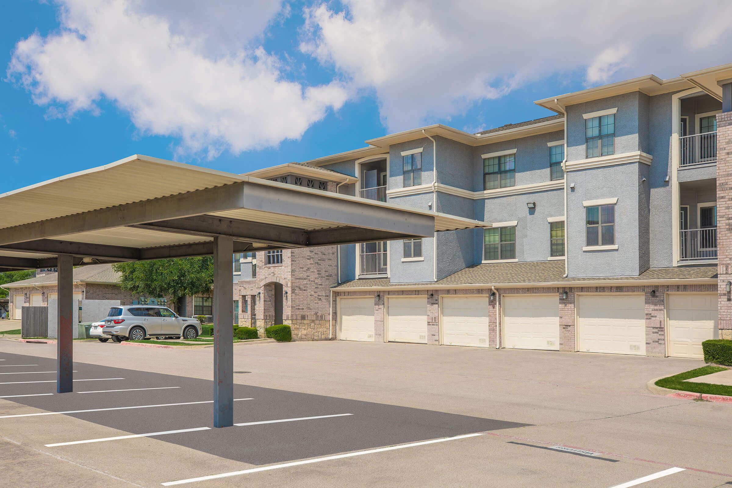 A parking lot with several empty spaces, featuring a covered area and a couple of parked cars. In the background, there are modern apartment buildings with light blue and beige exteriors under a partly cloudy sky.
