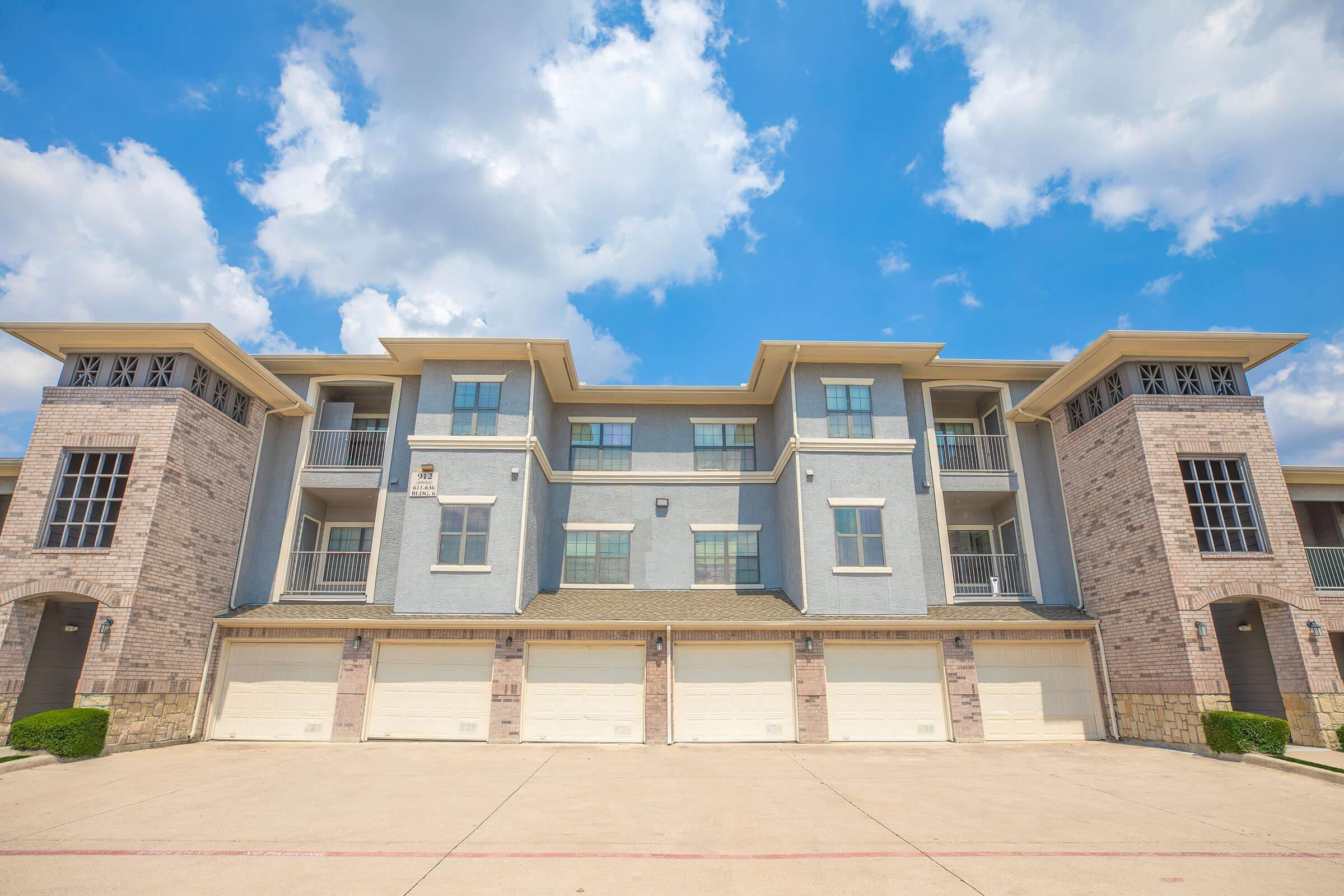 A modern apartment building with a symmetrical facade, featuring multiple balconies, large windows, and brick detailing. The sky is bright blue with fluffy white clouds, providing a sunny backdrop to the structure. The garages are visible at the bottom, and the overall design reflects contemporary architecture.
