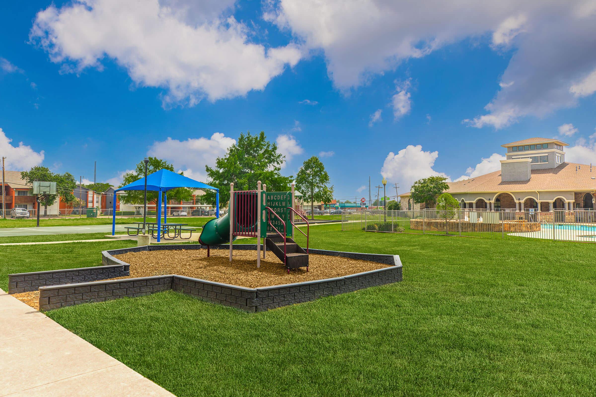 A vibrant playground area with a green slide and a climbing structure, surrounded by a grassy lawn. In the background, there is a swimming pool and a multi-story building with a decorative roof under a bright blue sky with scattered clouds. A shaded picnic area is nearby, along with a basketball court.