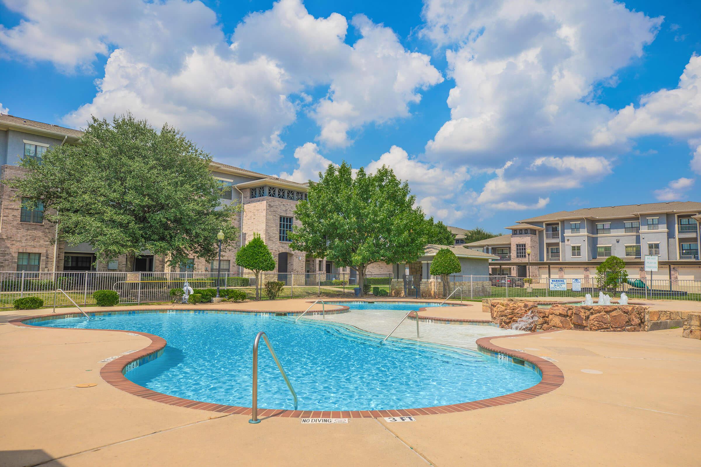 A spacious outdoor swimming pool surrounded by landscaped areas, featuring a gentle curve and accessible steps. In the background, there are residential buildings with large windows and balconies, under a bright blue sky with fluffy white clouds.