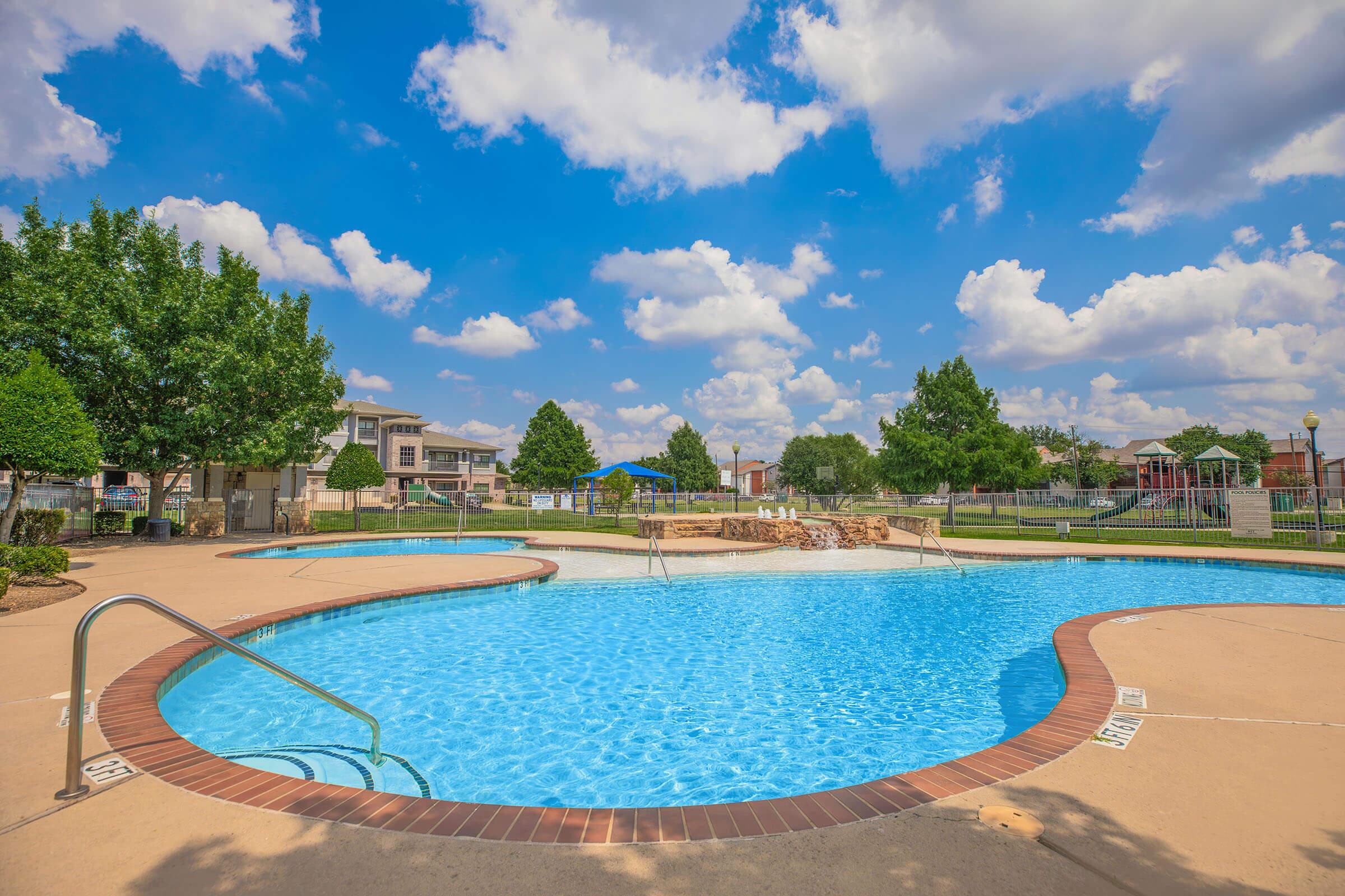 A sparkling blue swimming pool surrounded by a patio area. Lush green trees line the pool, and there are homes in the background. The sky is bright with fluffy white clouds, creating a cheerful and inviting atmosphere. A playground can be seen in the distance, adding to the recreational appeal of the area.
