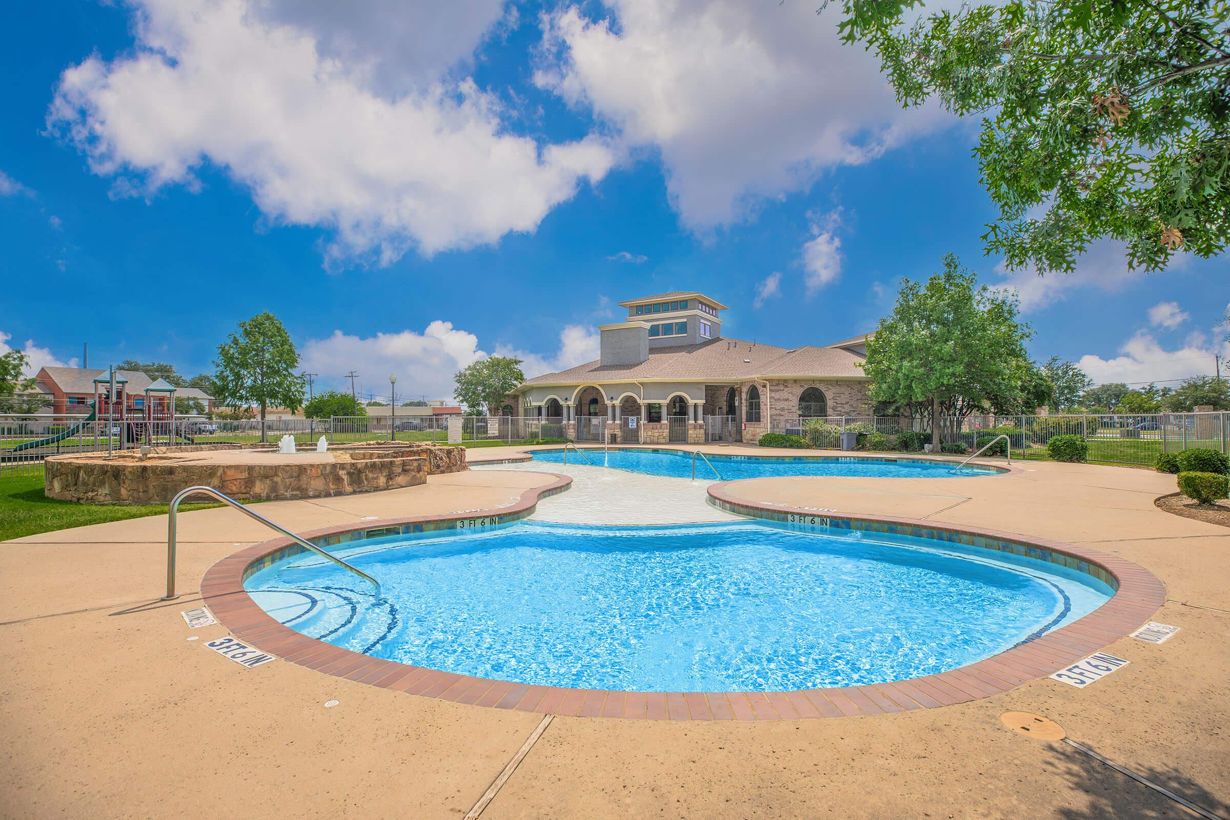 A vibrant outdoor pool area featuring two interconnected pools with clear blue water, surrounded by smooth concrete. Lush green trees line the vicinity, with a clubhouse visible in the background. A bright blue sky with fluffy white clouds adds to the scenic atmosphere. Suitable for relaxation and recreation.