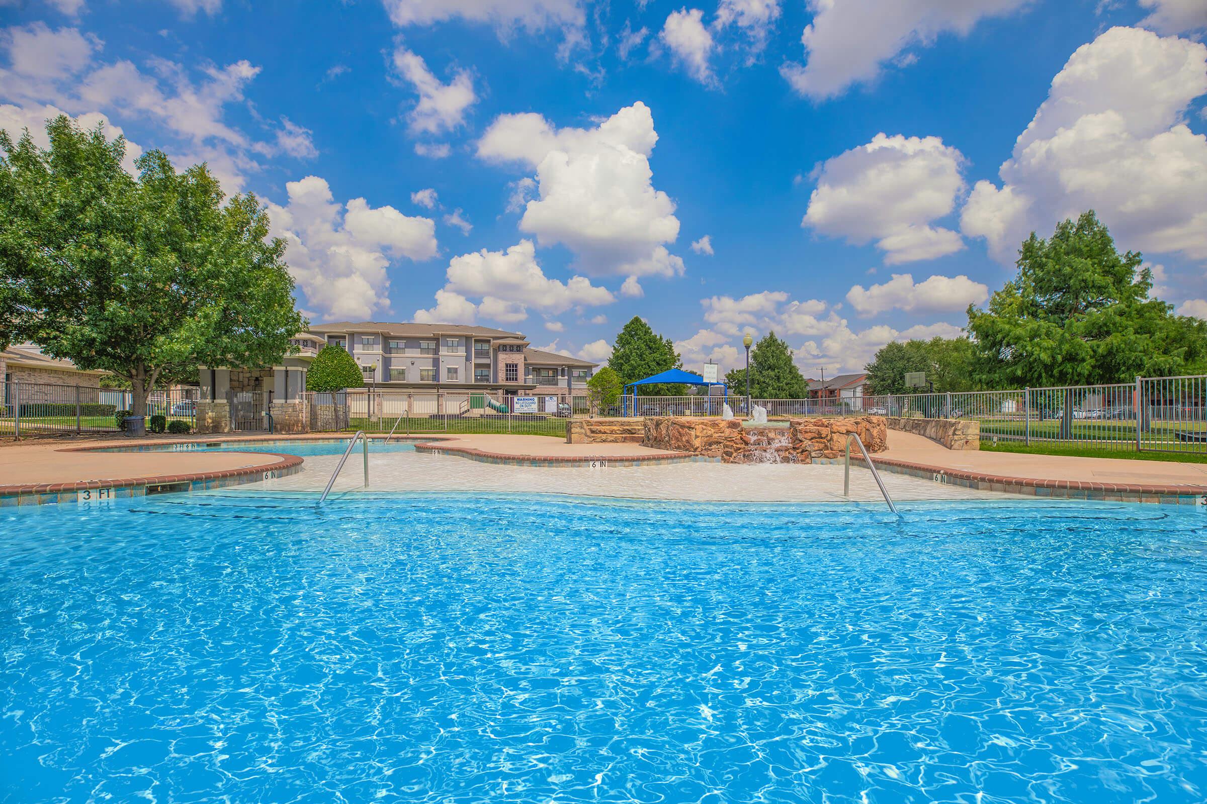 A sparkling blue swimming pool surrounded by lush green trees and residential buildings, under a bright blue sky dotted with fluffy white clouds. The tranquil scene invites relaxation and leisure, with the pool's clear water shimmering invitingly.