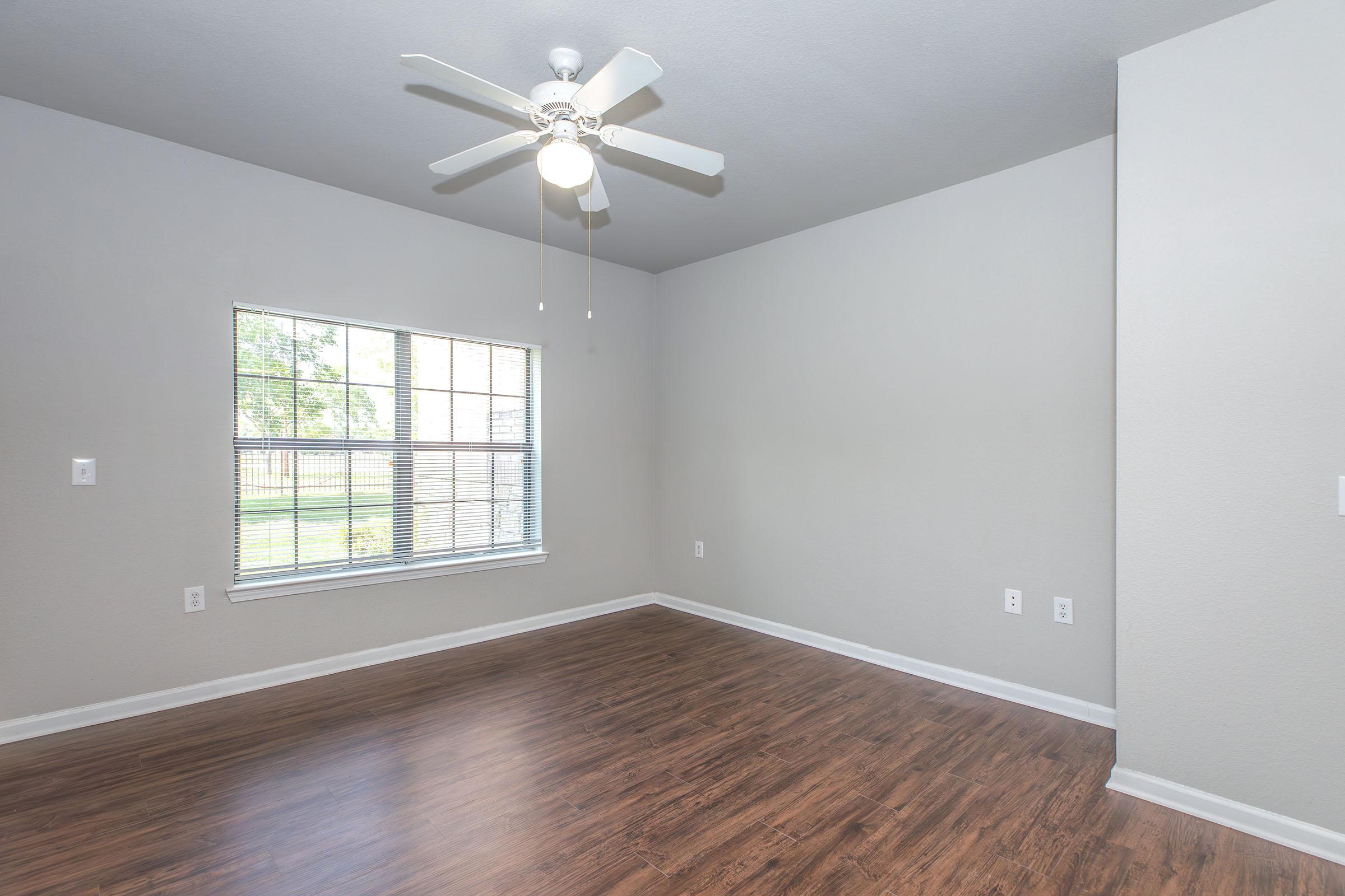 A spacious, unfurnished room with light gray walls and a wooden floor. A ceiling fan is mounted on the ceiling, and a window with multiple panes allows natural light to enter, casting a bright ambiance. The interior is minimalist, with no furniture present, creating an open and airy feel.