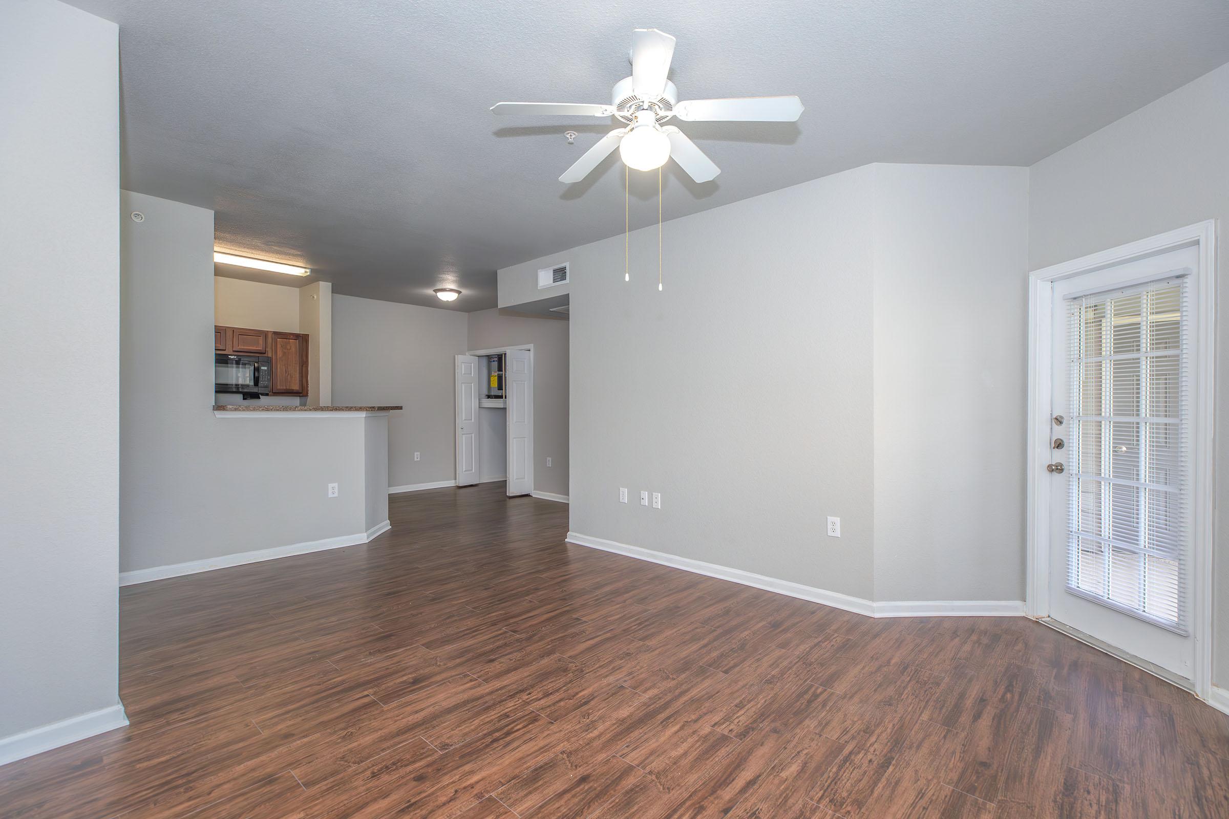 Empty living room with wood-style flooring, a ceiling fan, and neutral-colored walls. A partial view of a kitchen with wooden cabinets is visible in the background. A sliding glass door allows natural light and leads to an outdoor space. The layout is open and spacious.