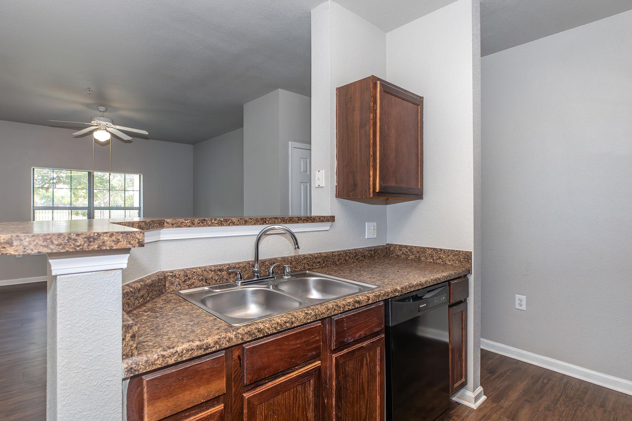 A modern kitchen area featuring a double sink, brown granite countertop, and dark wood cabinetry. The view includes a living space with light walls and a ceiling fan, and a window allowing natural light to enter the room. A black dishwasher is visible beside the sink.