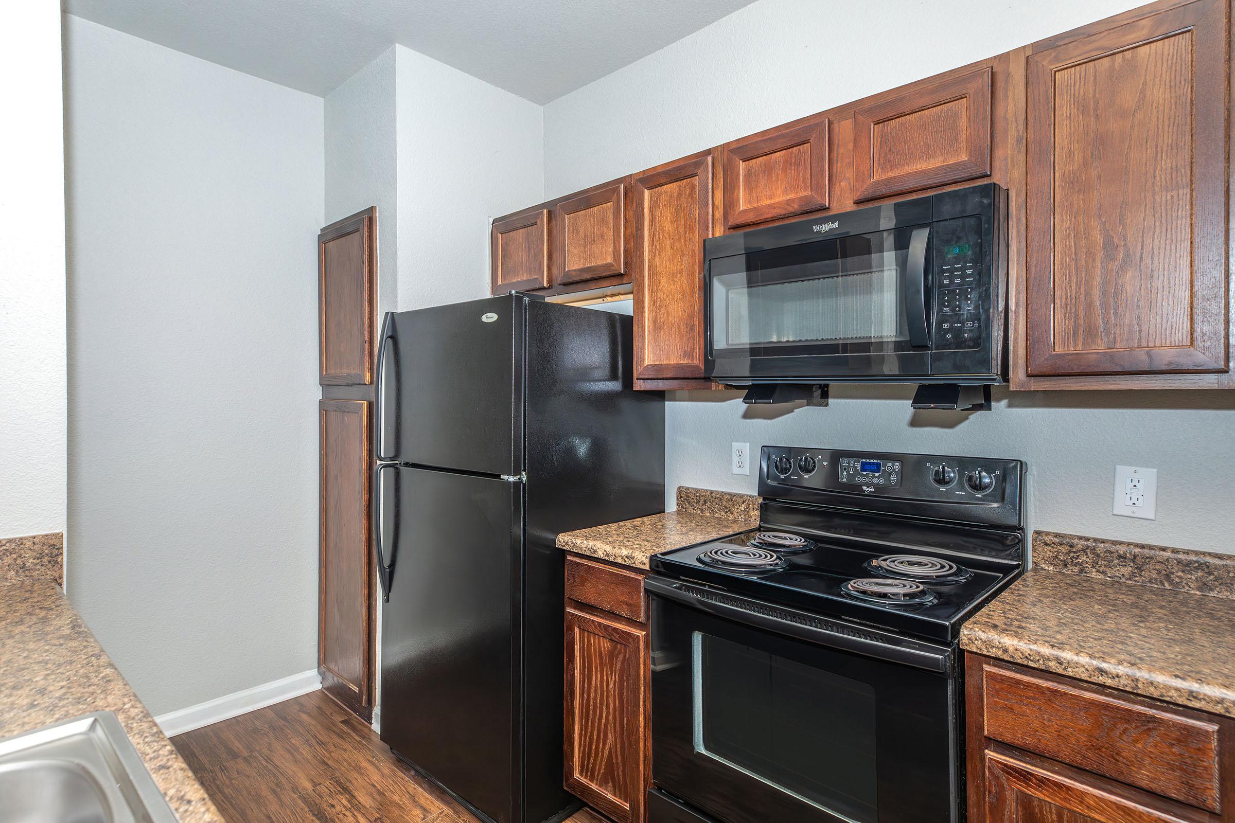 A modern kitchen featuring dark wood cabinetry, a black refrigerator, a built-in microwave above a smooth-top stove, and a countertop with a textured design. The walls are painted in a light color, and the flooring is dark to contrast with the cabinetry.