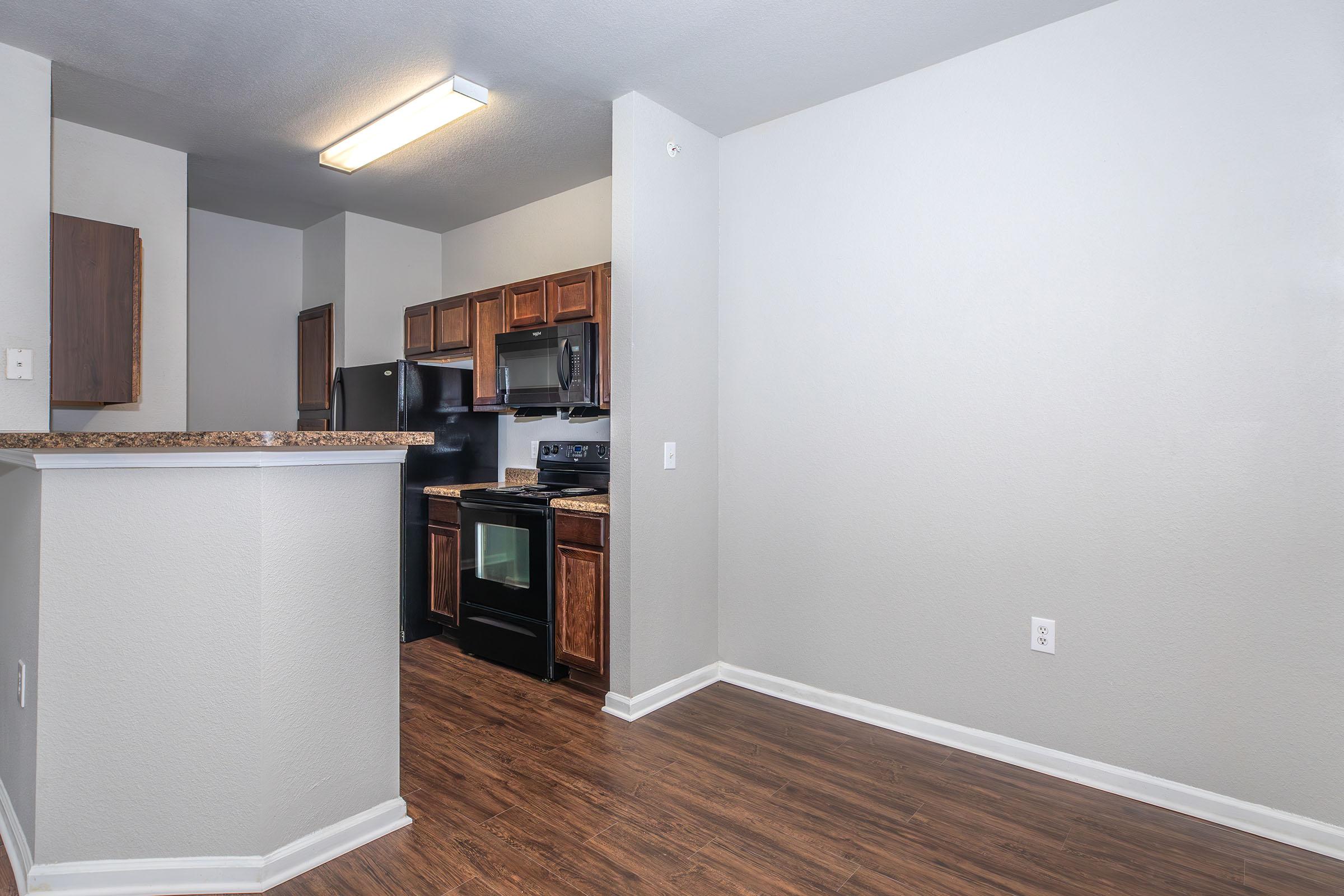 A modern kitchen with dark wooden cabinets, black appliances, and a granite countertop. The space features laminate flooring and light-colored walls, creating a bright and open atmosphere. A small bar area separates the kitchen from the living space, enhancing the functional layout.