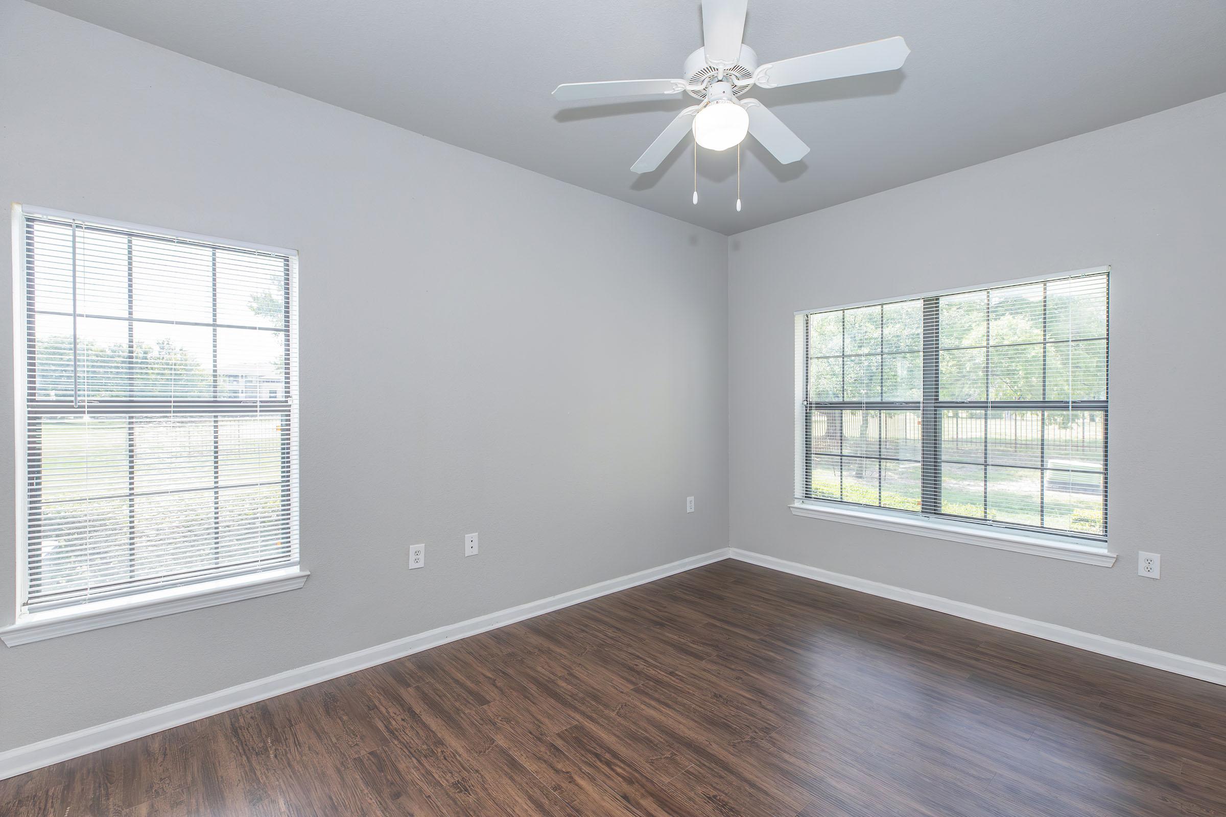 A well-lit room with light-colored walls and a wooden floor. Two large windows with white blinds allow natural light to enter. A ceiling fan is visible above, enhancing the spacious feel of the area. The room is empty, providing a blank canvas for decoration or furniture placement.