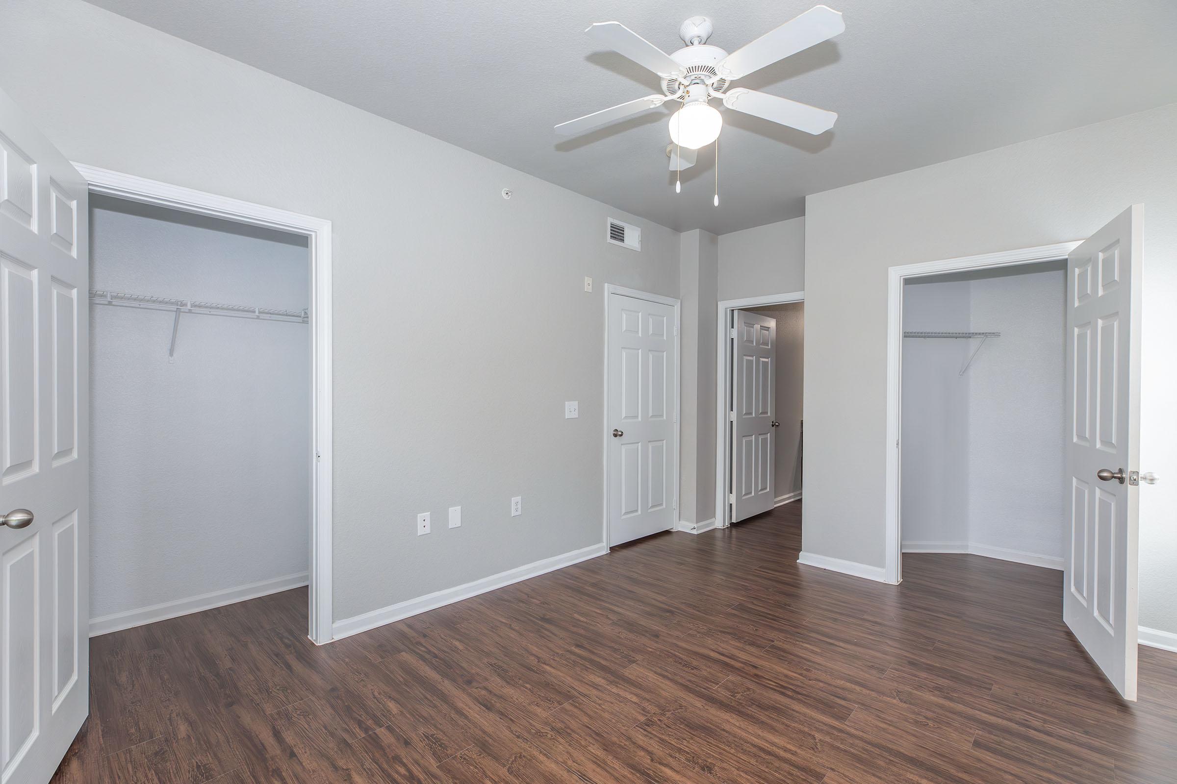 A bright, empty room with light gray walls and dark wood flooring. There are two open doors leading to closets on the left and right sides. A ceiling fan is visible above, providing a modern touch to the space. Natural light fills the area, creating a cheerful and spacious atmosphere.