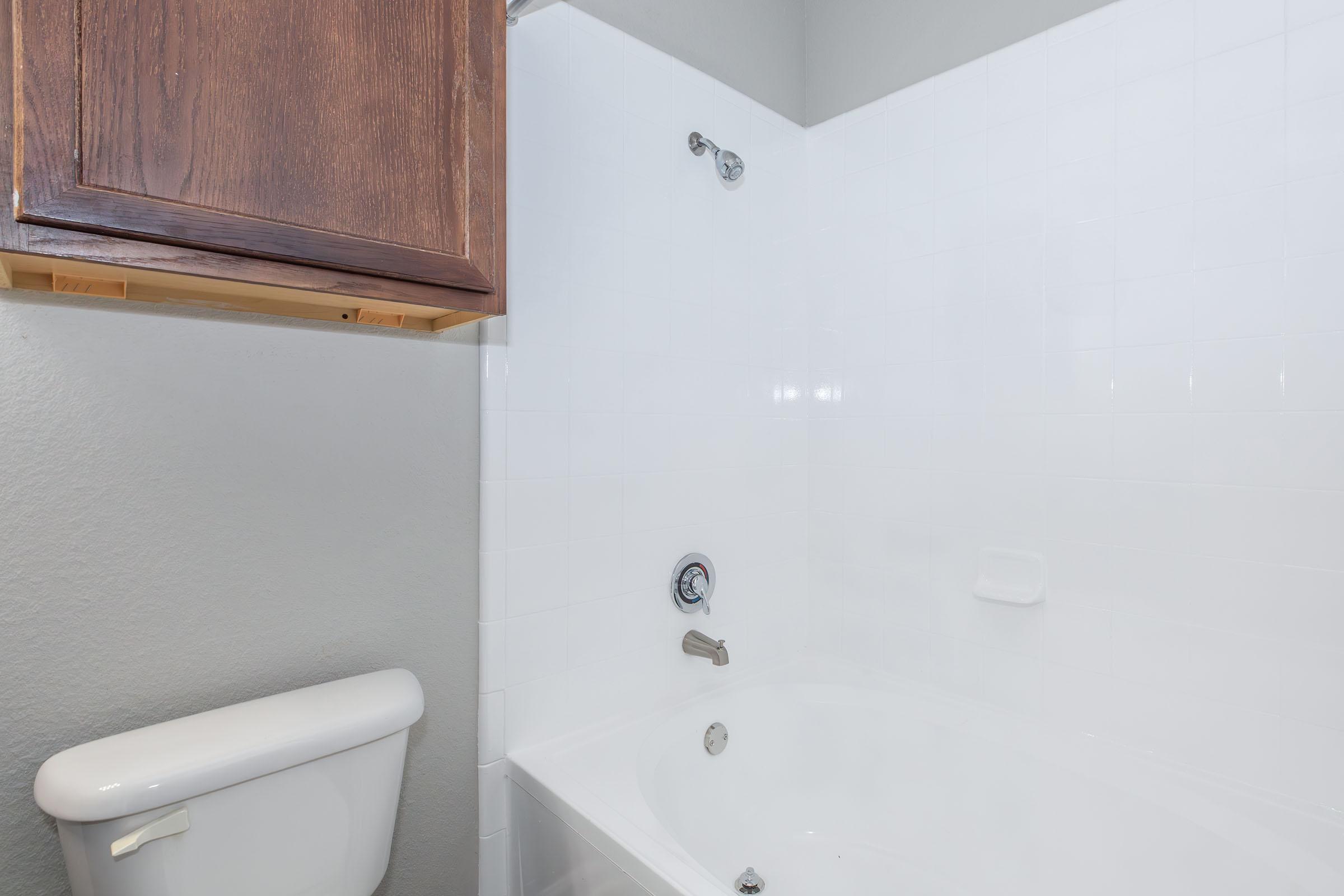 A clean, modern bathroom featuring a white tiled tub and shower with a chrome faucet, a white toilet nearby, and a wooden cabinet above the tub. The walls are painted a light gray, creating a bright and spacious atmosphere.
