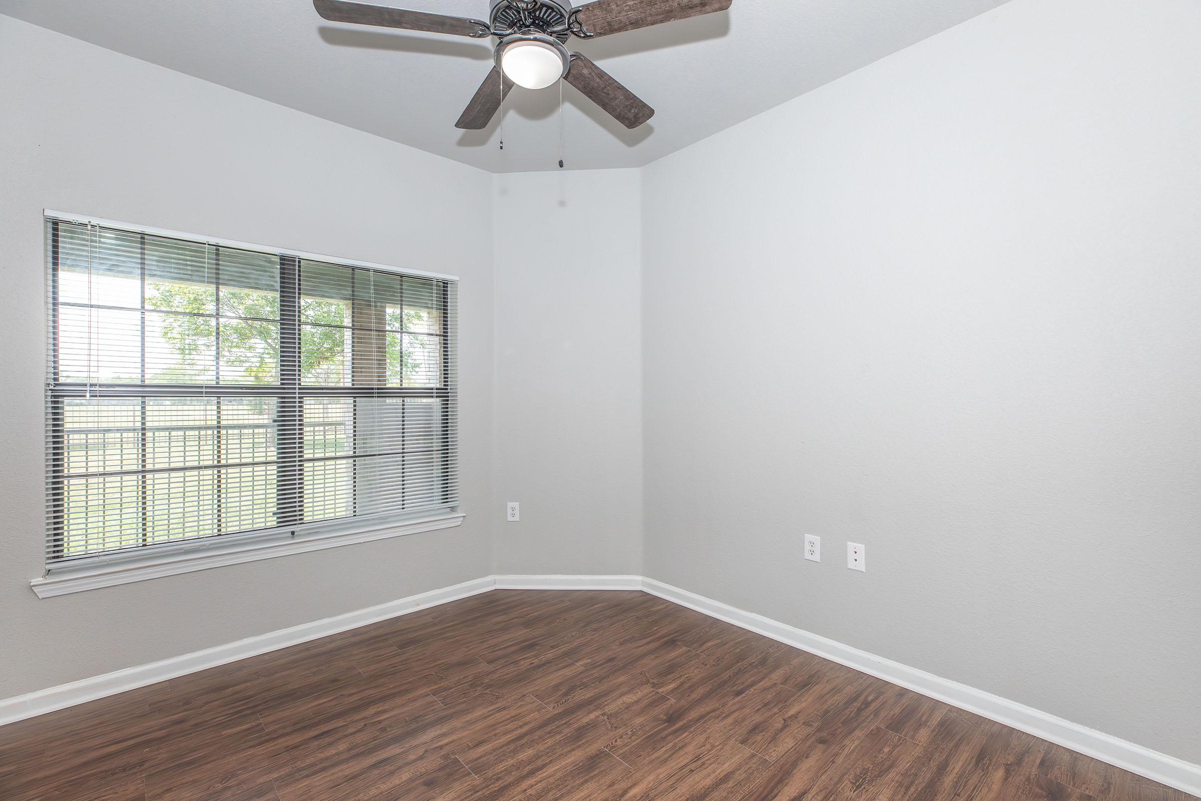 A clean, empty room with light gray walls and dark wood laminate flooring. There is a ceiling fan with wooden blades, and a window with blinds that lets in natural light, revealing a view of greenery outside. The room has two wall sockets and is ready for furniture placement.