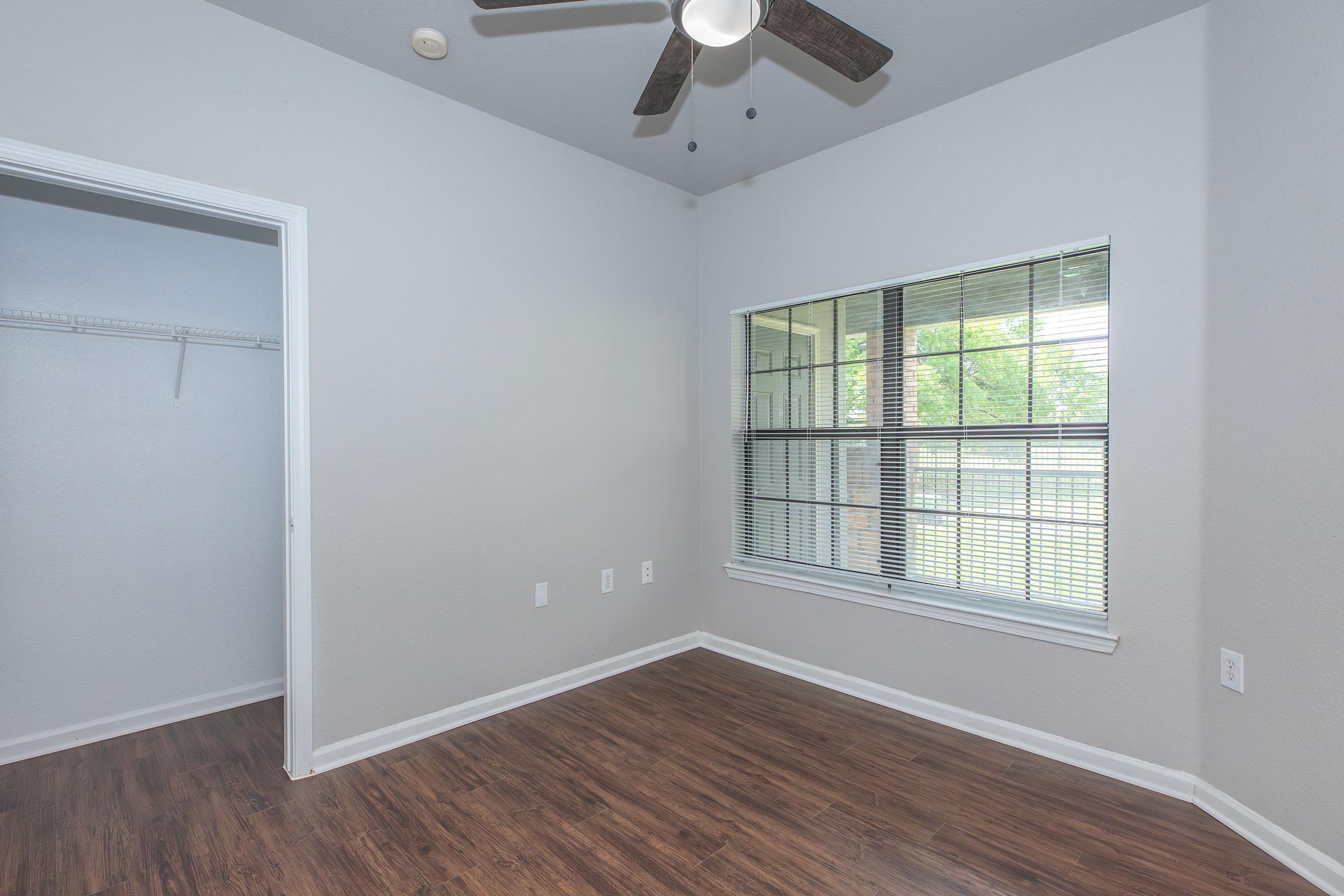 Empty room with light gray walls and a wooden floor. A window with blinds lets in natural light, and there is a ceiling fan visible. A doorway leads to a closet with a hanging rack. The overall space is bright and minimalistic, suitable for various purposes such as a bedroom or office.