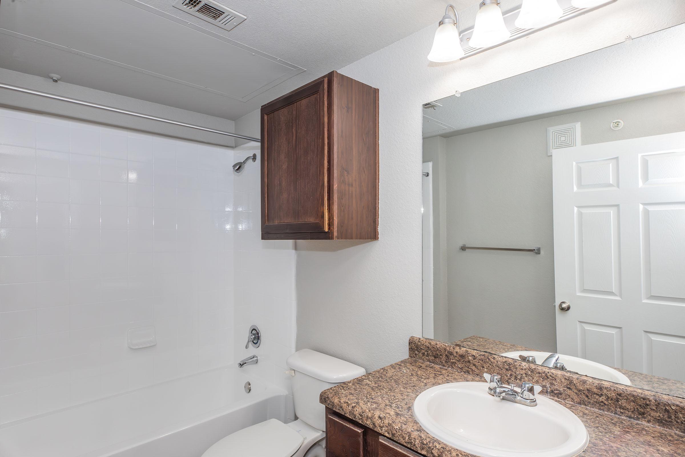 A bathroom featuring a white bathtub with a shower, a dark wooden cabinet above the sink, a beige countertop, and a mirror. The walls are painted light gray, and there is a towel rack to the right. Bright lighting is provided by three fixtures above the mirror.