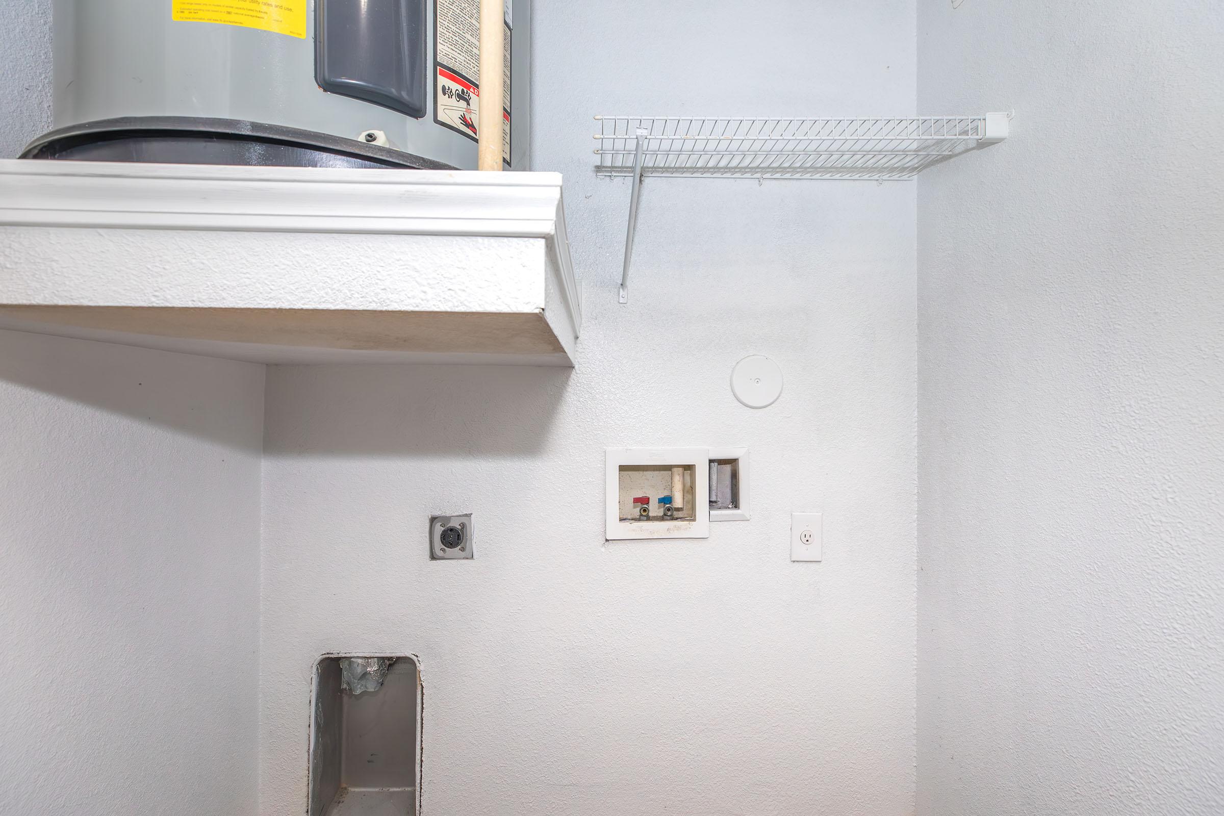 A small, empty laundry area featuring a water heater mounted on the wall, a shelf above, and a wall with electrical connections. There is a wire rack mounted on the wall for storage. The space has plain, light-colored walls and a visible, unfinished section where plumbing is installed.