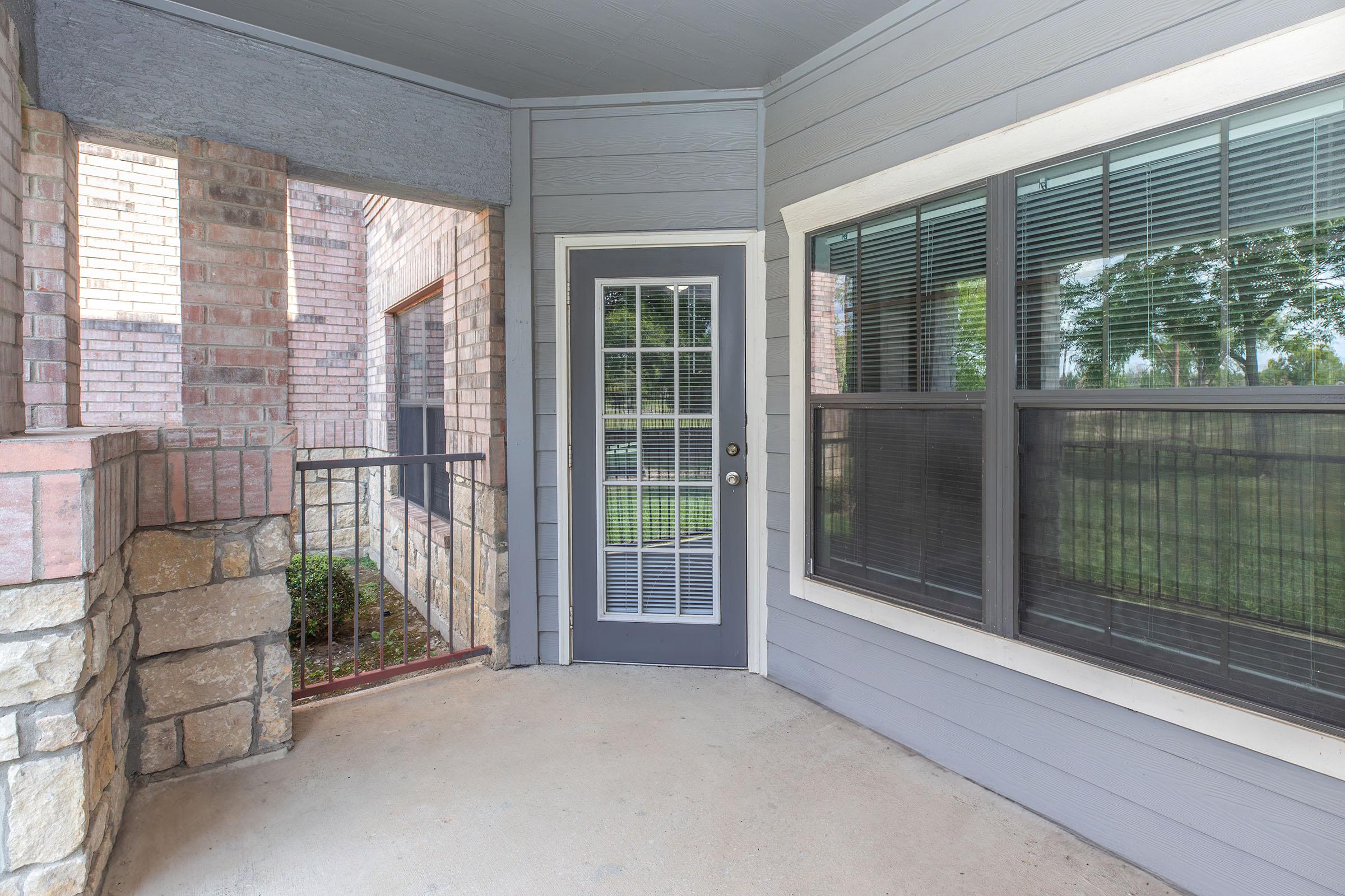 A corner view of a porch featuring a gray door with vertical glass panels, surrounded by light gray and brick walls. Large windows with grid patterns allow natural light. The porch has a concrete floor and a railing, with greenery visible outside.