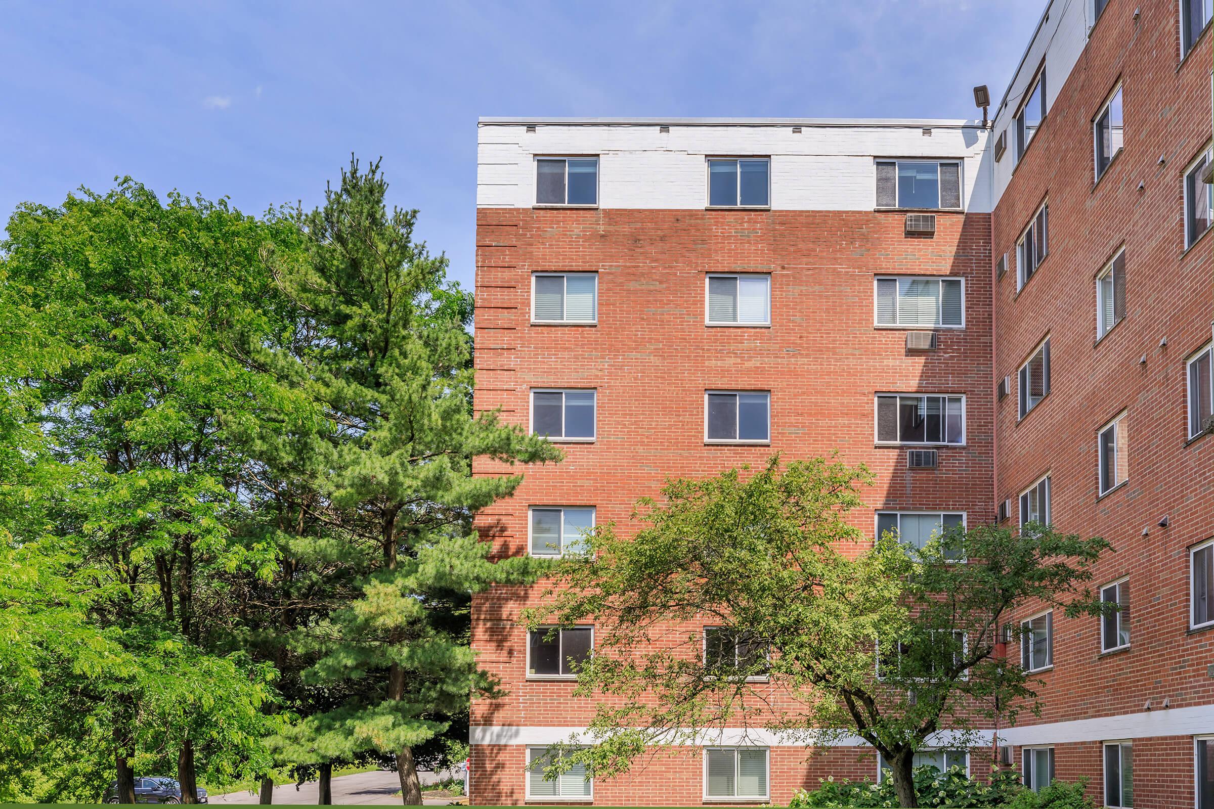A brick apartment building with multiple windows is partially visible. The exterior features a reddish-brown color, and it's surrounded by trees, with a clear blue sky in the background. The scene reflects a residential area with greenery.