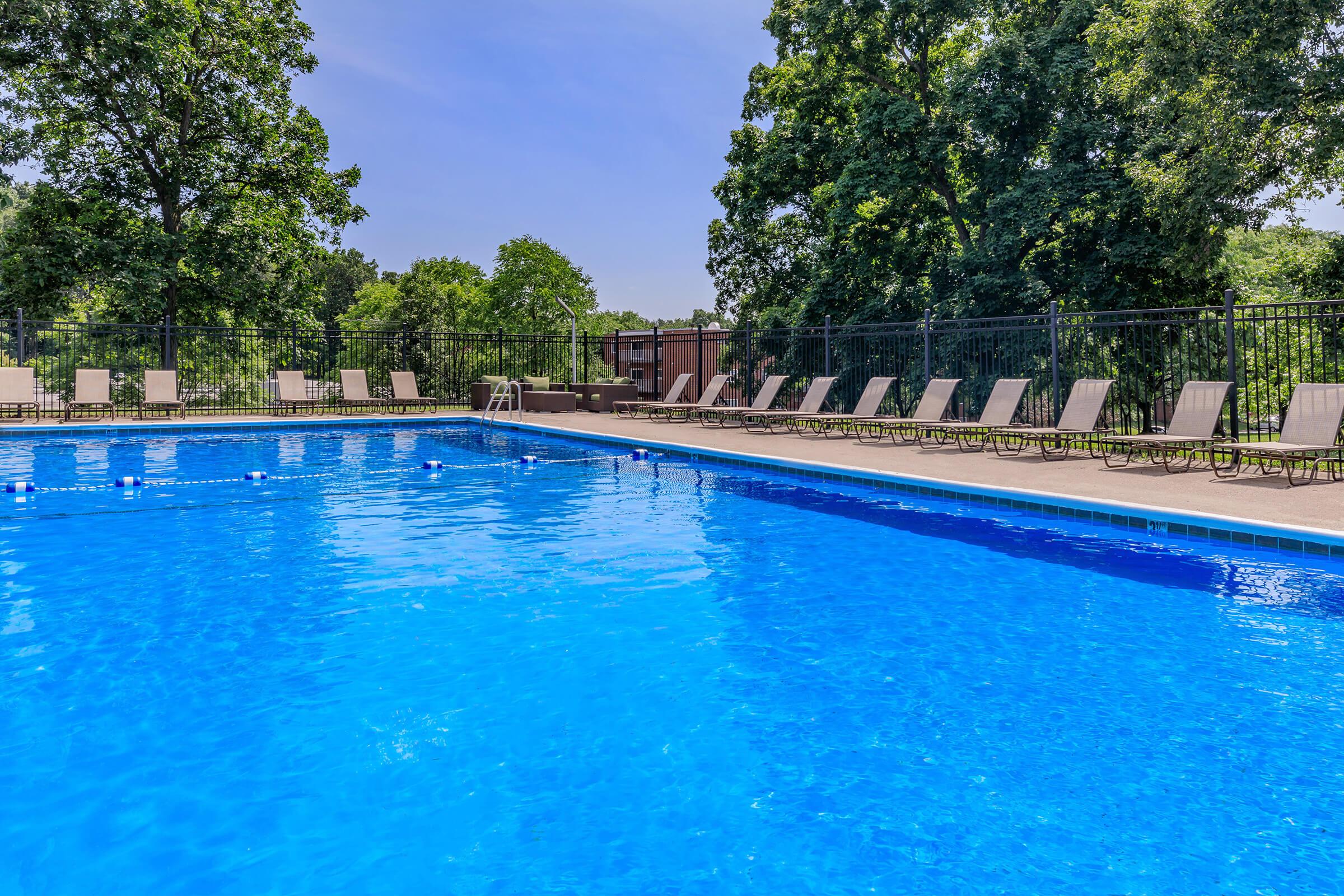 A clear blue swimming pool surrounded by green trees, with several lounge chairs lined up along the poolside. The sky is bright and sunny, creating a relaxing outdoor atmosphere perfect for leisure and enjoyment.