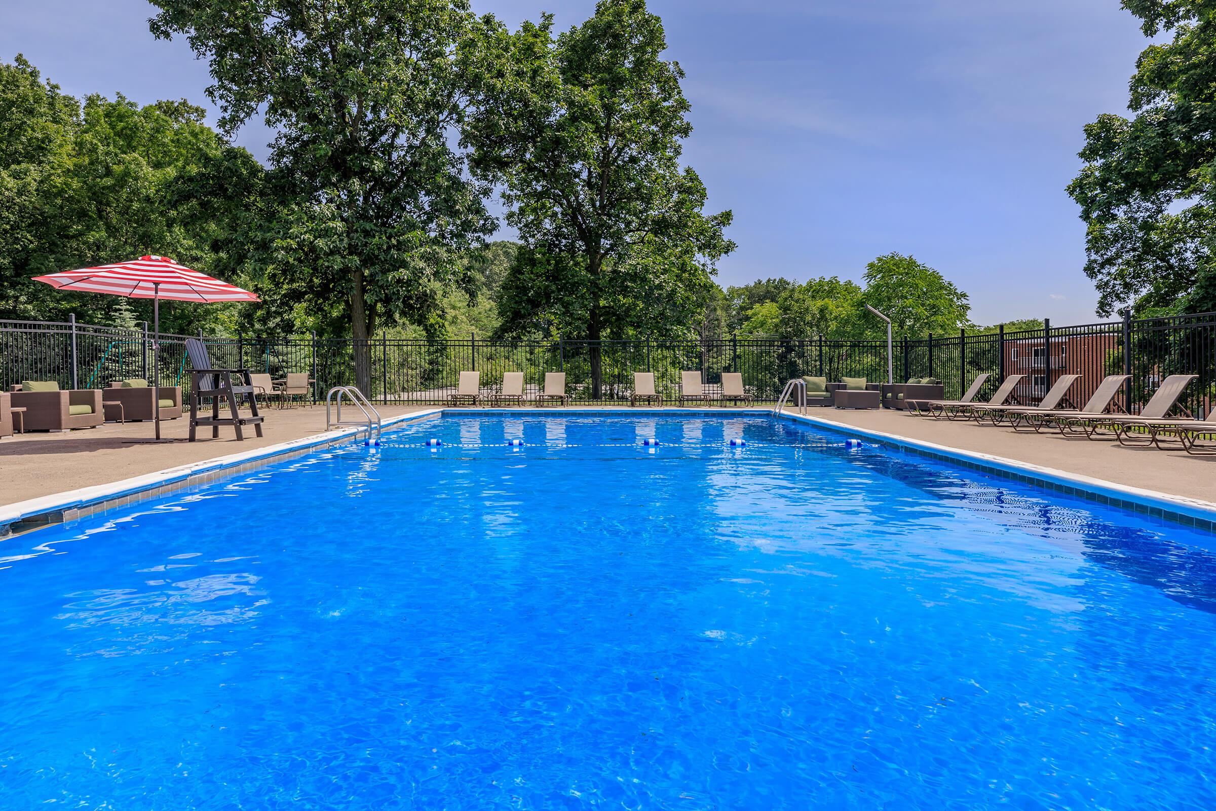 A serene outdoor swimming pool with clear blue water, surrounded by lush green trees. Sun loungers line the pool area, and a red and white striped umbrella provides shade. The sky is clear and blue, creating a picturesque summer setting.