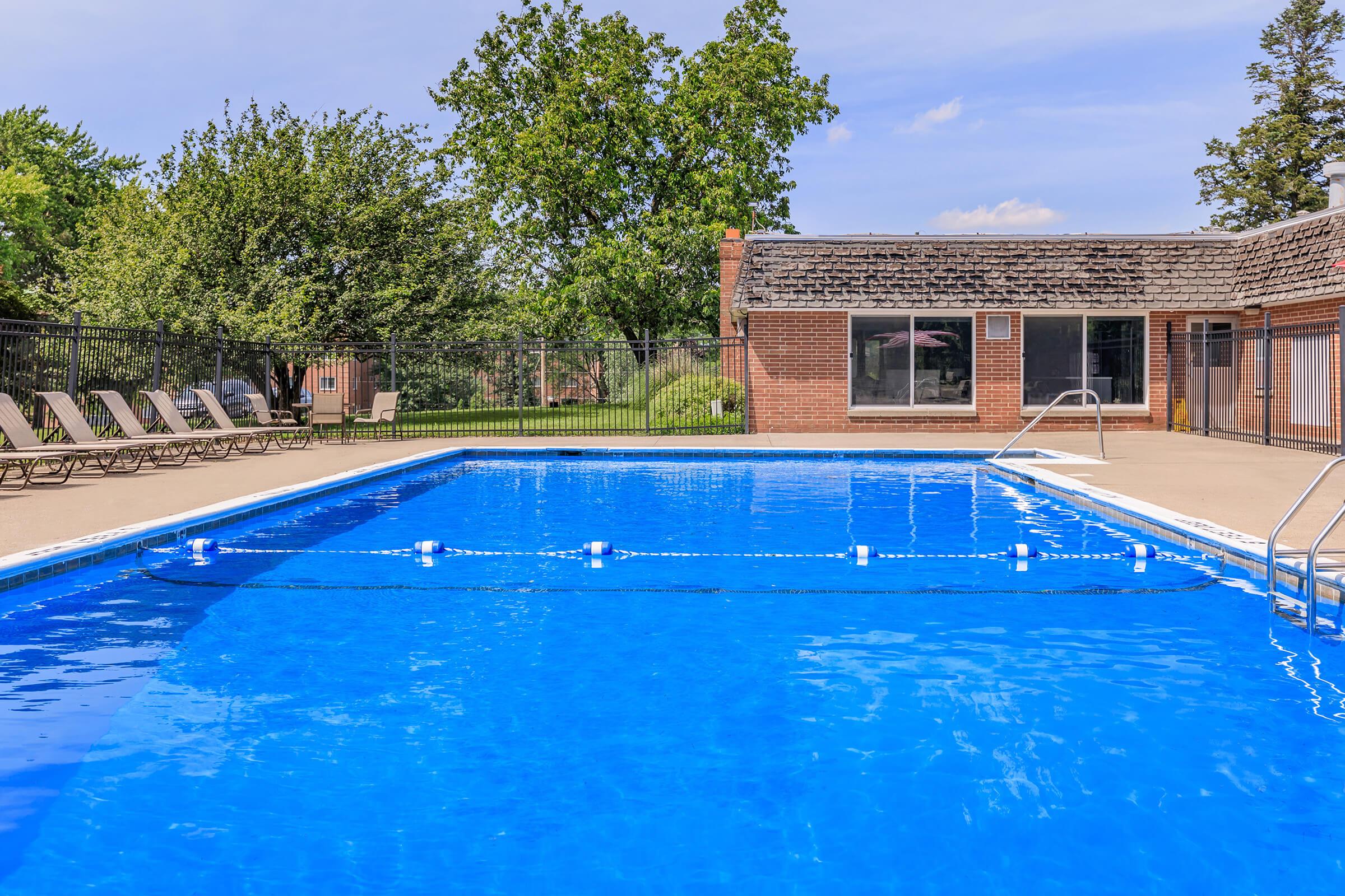 A clear blue swimming pool with lounge chairs on the surrounding deck. Lush green trees and grass can be seen in the background, along with a brick building featuring large windows. The atmosphere is bright and inviting, suggesting a relaxing outdoor space.