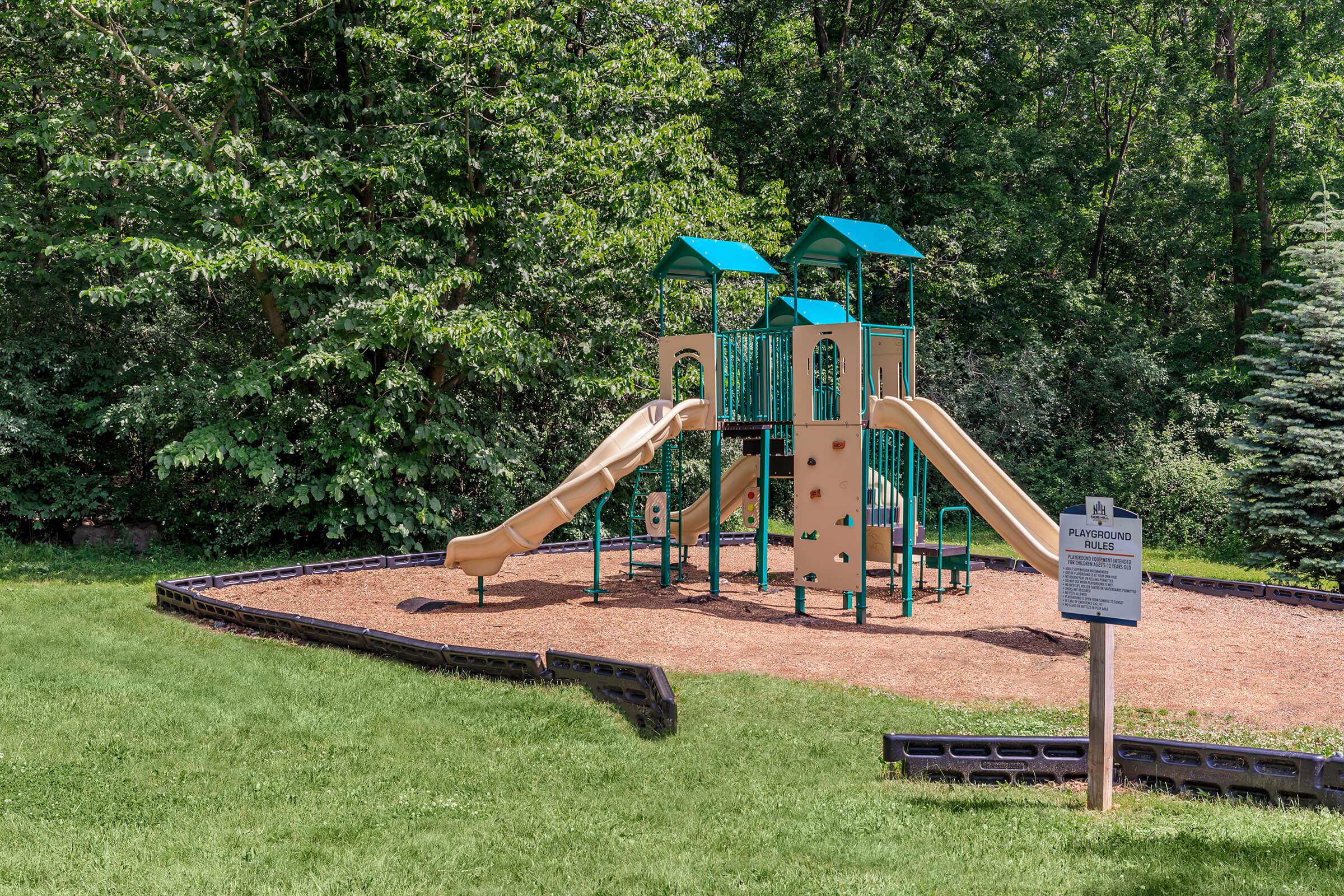 A colorful playground featuring two slides, climbing structures, and a climbing wall surrounded by green grass and trees. A sign indicating playground rules is visible in the foreground.